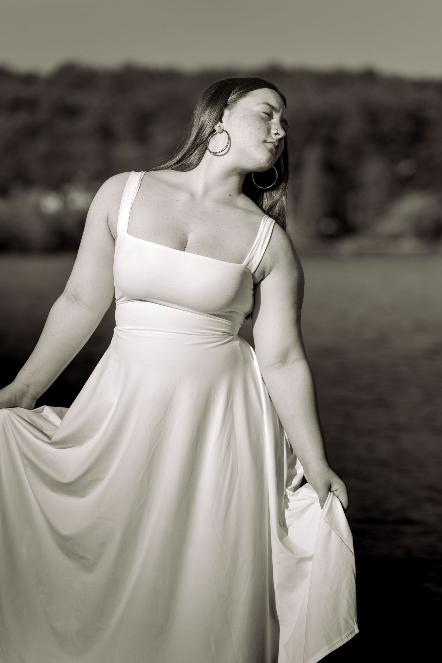 Black and white photo of a woman in a white dress posing outdoors near water with trees in background.