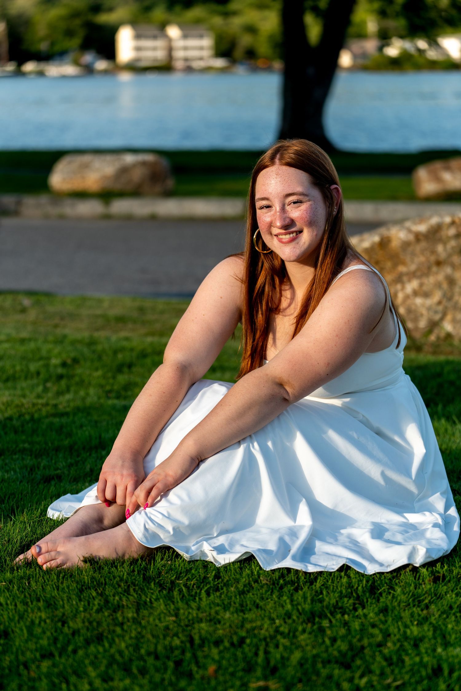 Young woman with red hair in white dress sitting on green grass by a lake, smiling in golden hour light.