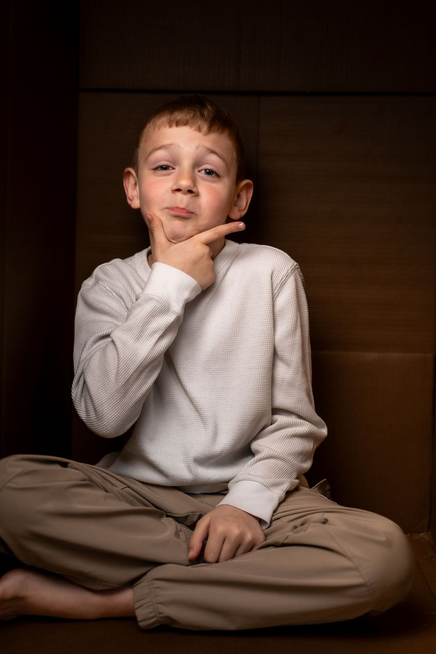 Young boy sitting cross-legged in a cardboard box, resting chin on hand, wearing white sweater and tan pants.