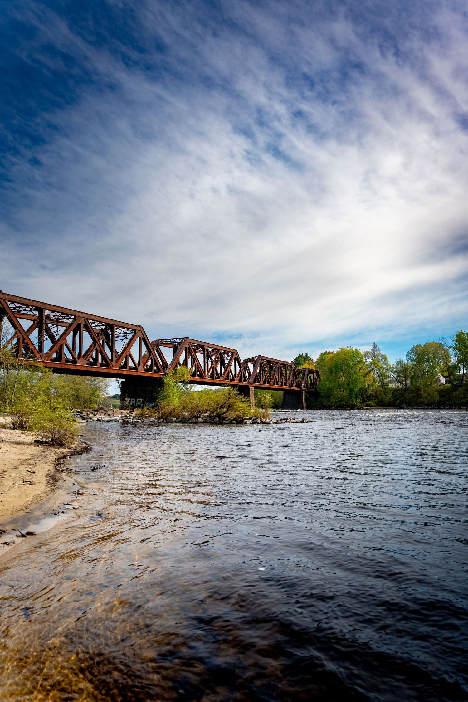 Rustic iron railway bridge spanning a calm river with sandy shores, lush trees, and dramatic cloudy blue sky.