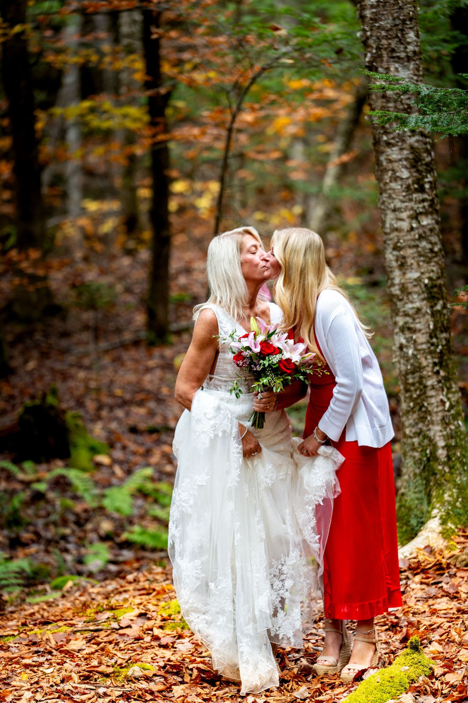 Two people in white and red dresses share tender moments among autumn leaves in a forest setting.