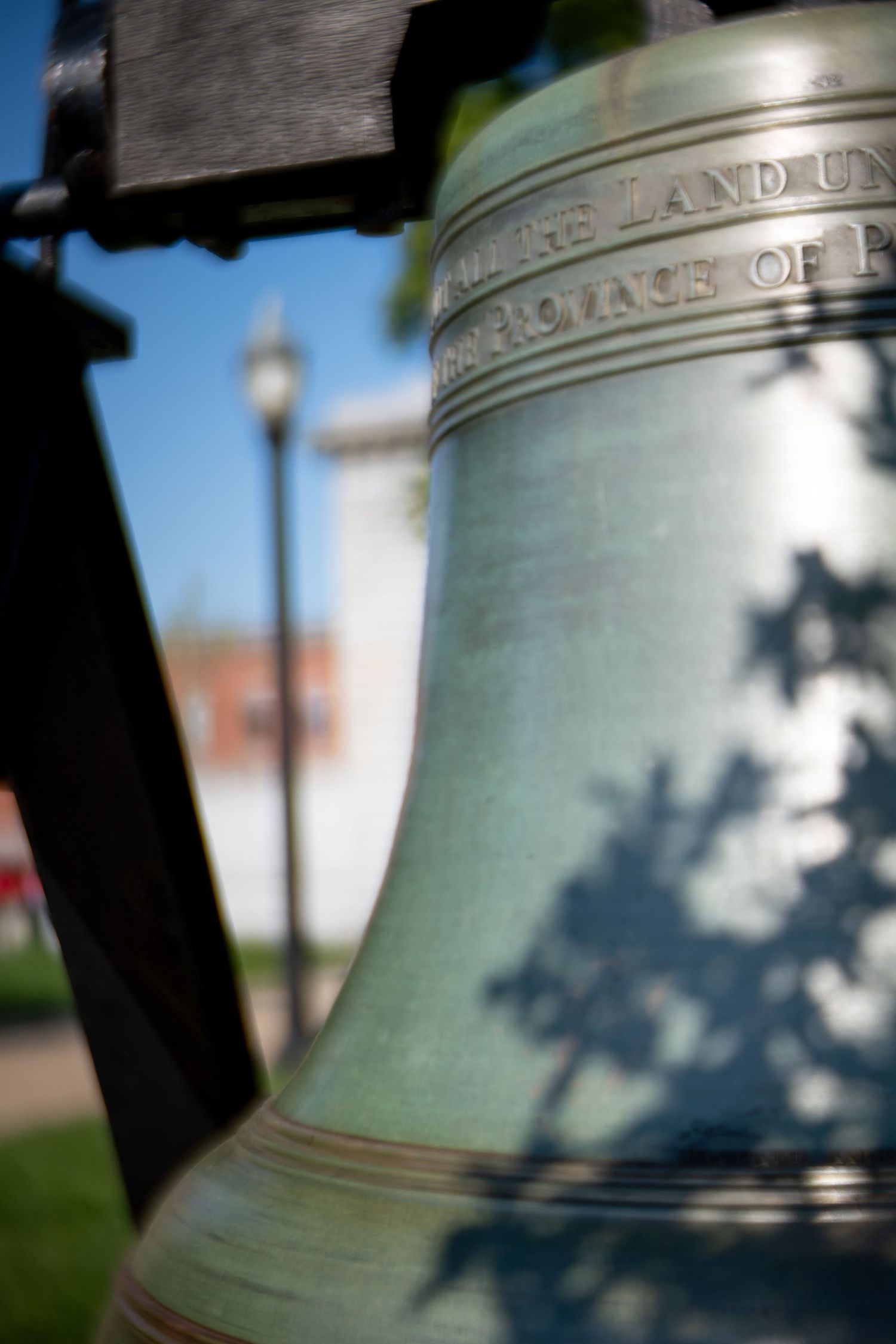 Close-up of a green patina Liberty Bell replica with inscribed text, mounted outdoors against a blue sky.