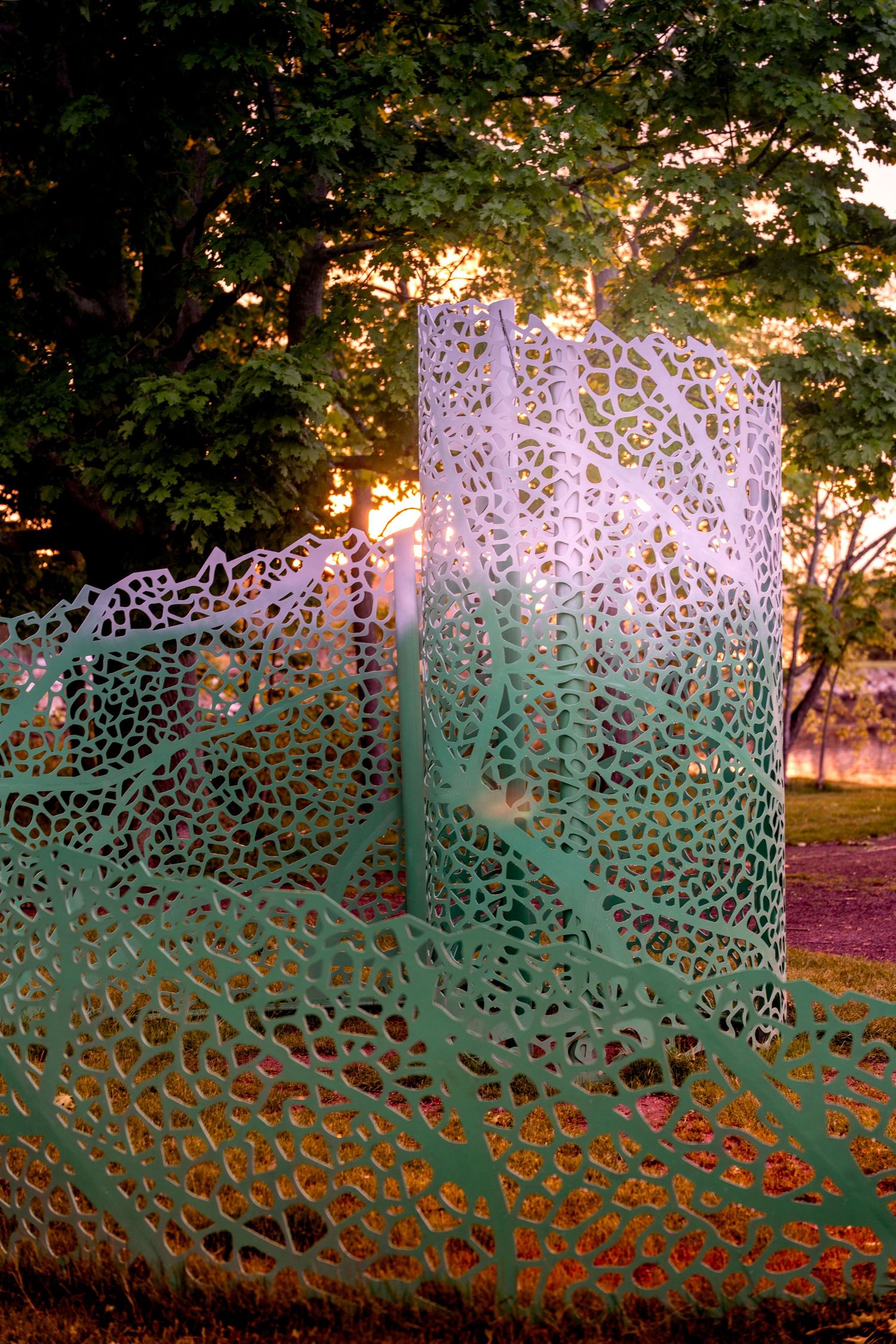 Intricate green metal lattice sculpture with organic patterns glowing at sunset in a lush park setting.