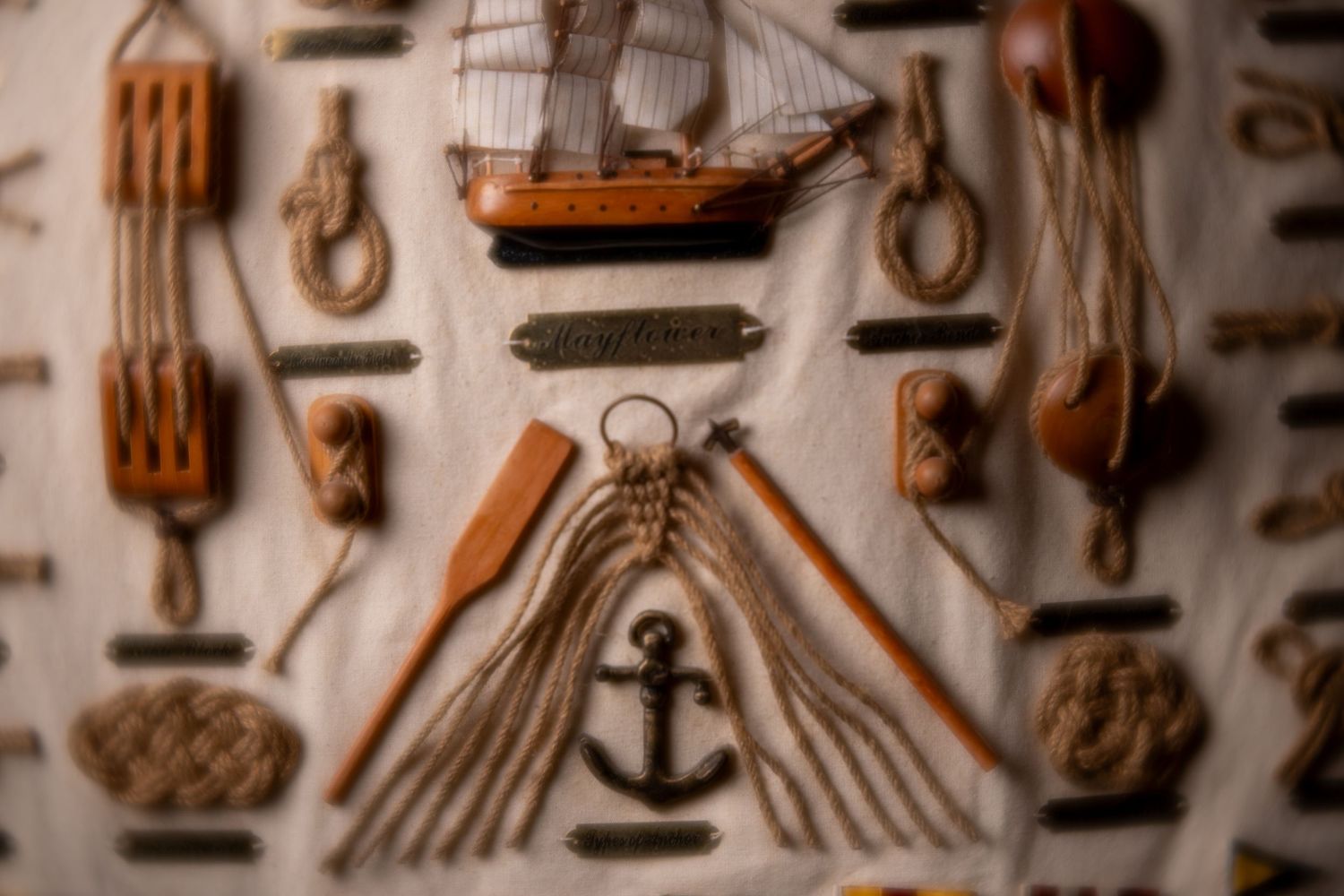 Nautical flat lay display featuring ship model, anchor, rope knots, wooden pulleys, and sailing tools on white fabric.