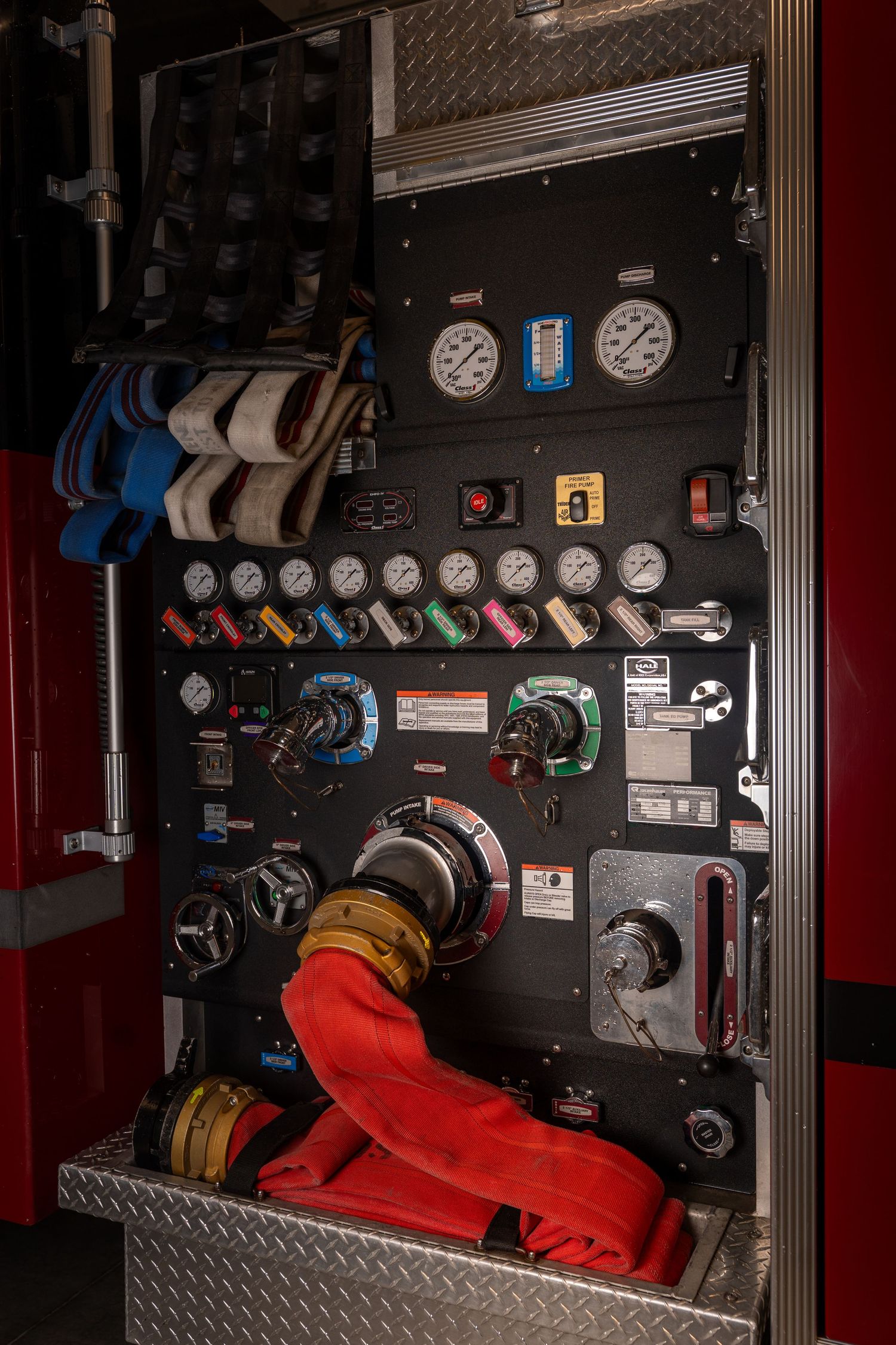 Fire truck pump panel with multiple pressure gauges, valves, and red hose connections.