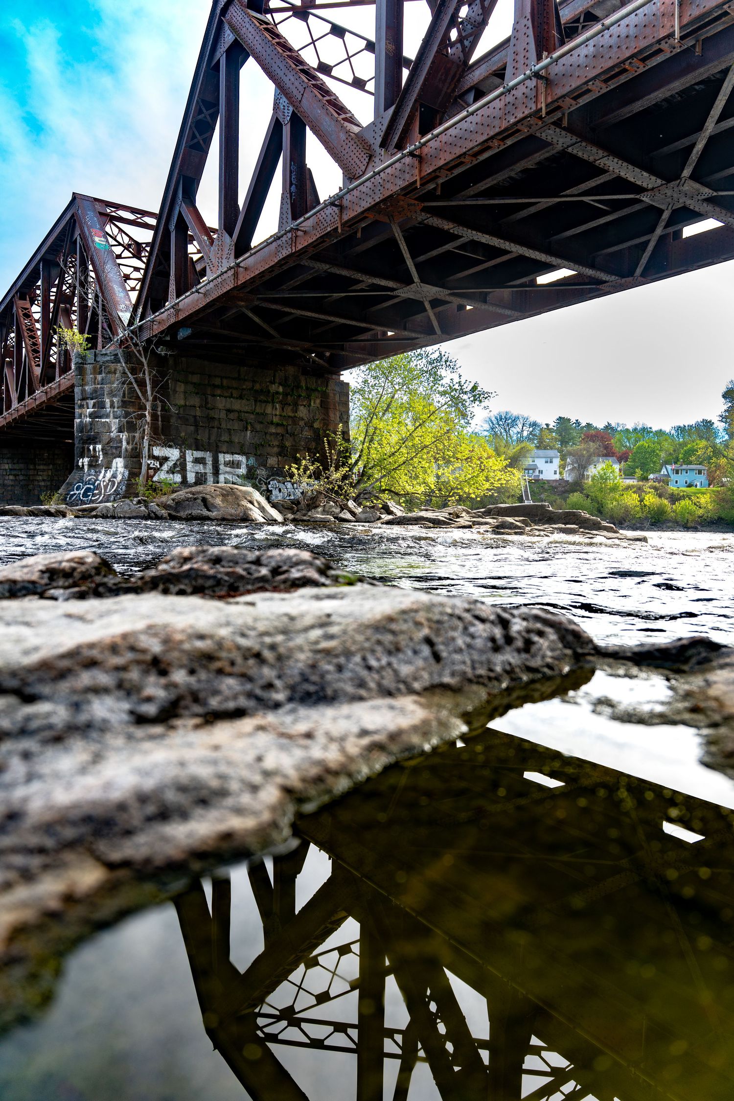 Rusty iron railroad bridge over rocky river, low angle view showing reflection in calm water with graffiti on stone pillar.