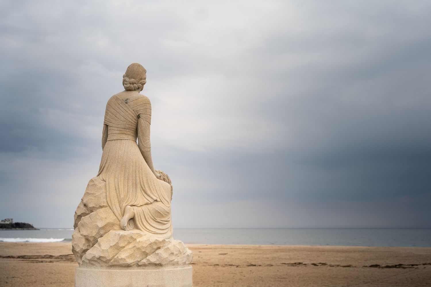 Stone statue of a seated woman facing the ocean on a sandy beach under a cloudy sky.
