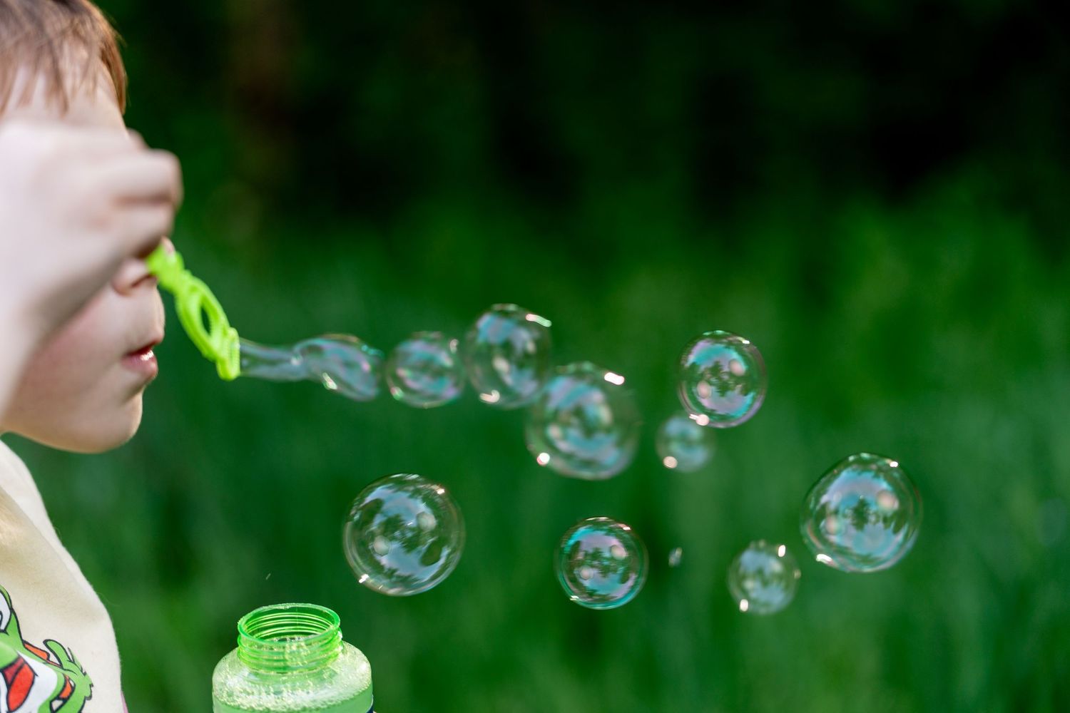 Child blowing soap bubbles outdoors with green bubble wand and bottle, bubbles floating in front of lush green grass.