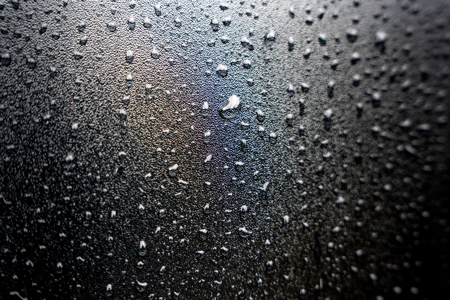 Close-up of water droplets on a dark gray surface with subtle light reflections creating a moody abstract texture.