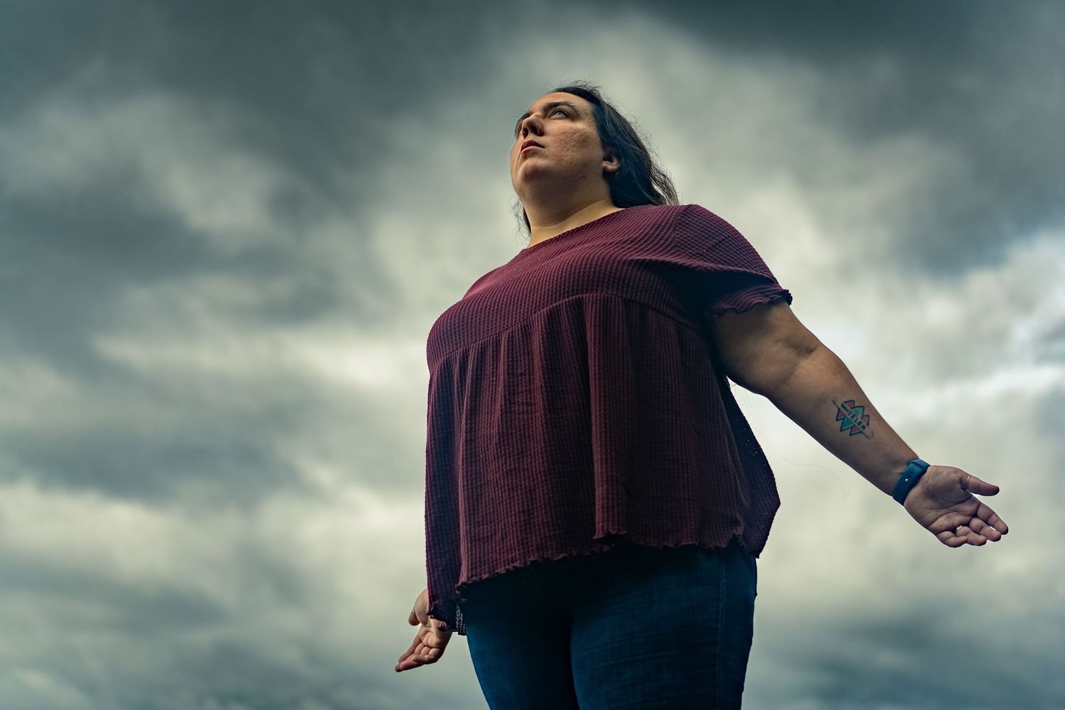 Person in burgundy shirt stands against dramatic cloudy sky with arms outstretched in emotional pose.