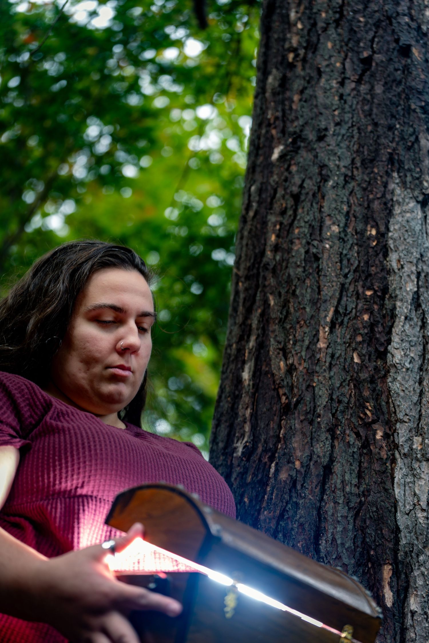 Person in red plaid shirt leans against tree trunk while holding glowing light stick in forest setting.