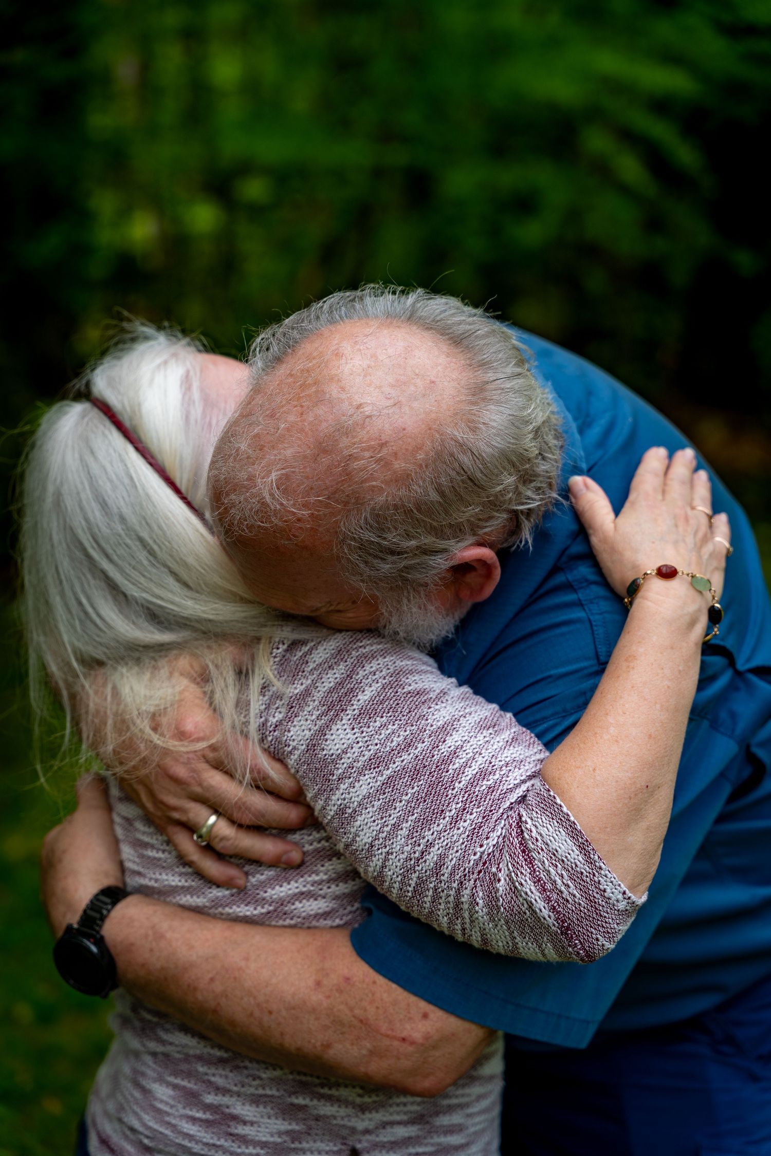 An elderly couple embraces in a tender hug outdoors against a green background.