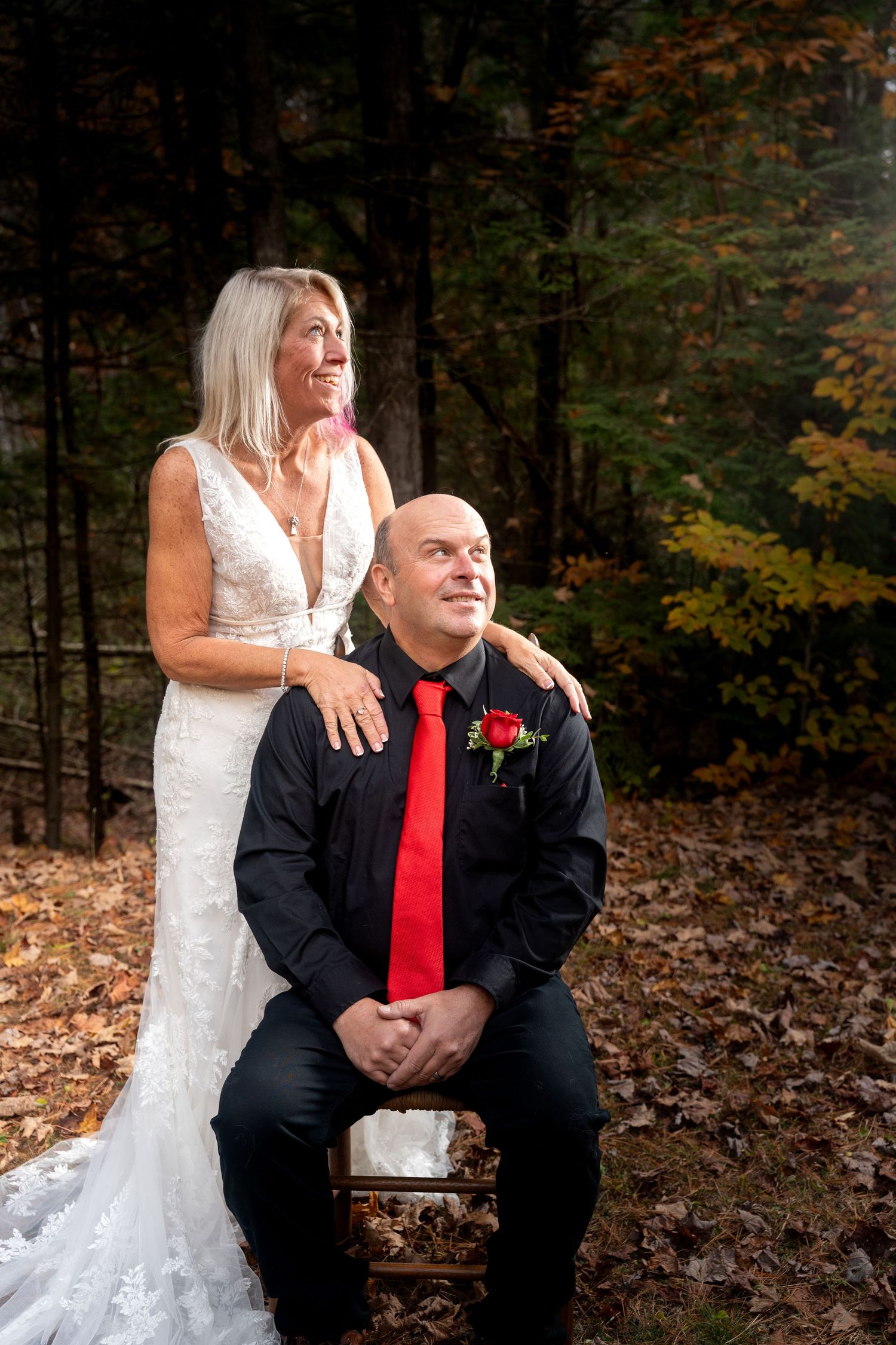 A couple in wedding attire poses together on a bench in a wooded autumn setting with fallen leaves.