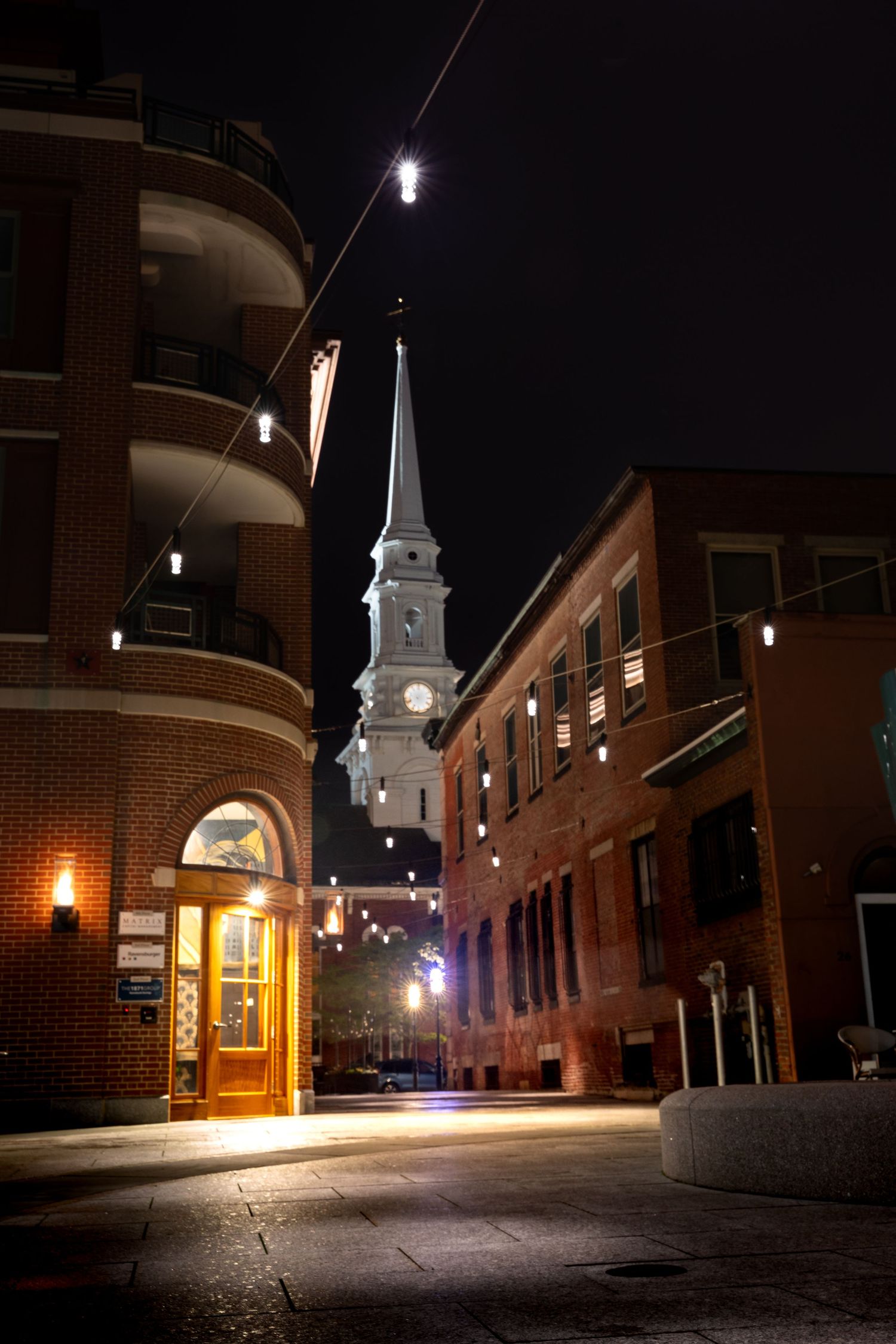 Night view of a historic church steeple rising above brick buildings on a quiet cobblestone street.