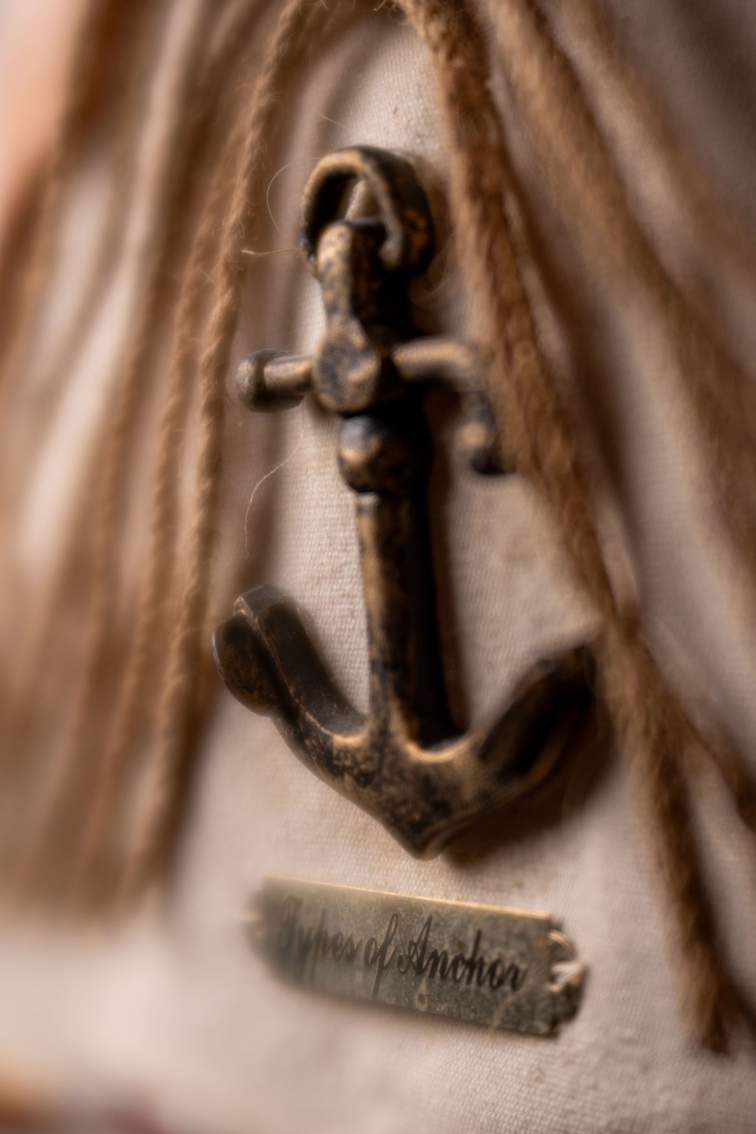 Vintage bronze anchor charm with rope and small metal tag reading types of anchor on fabric.