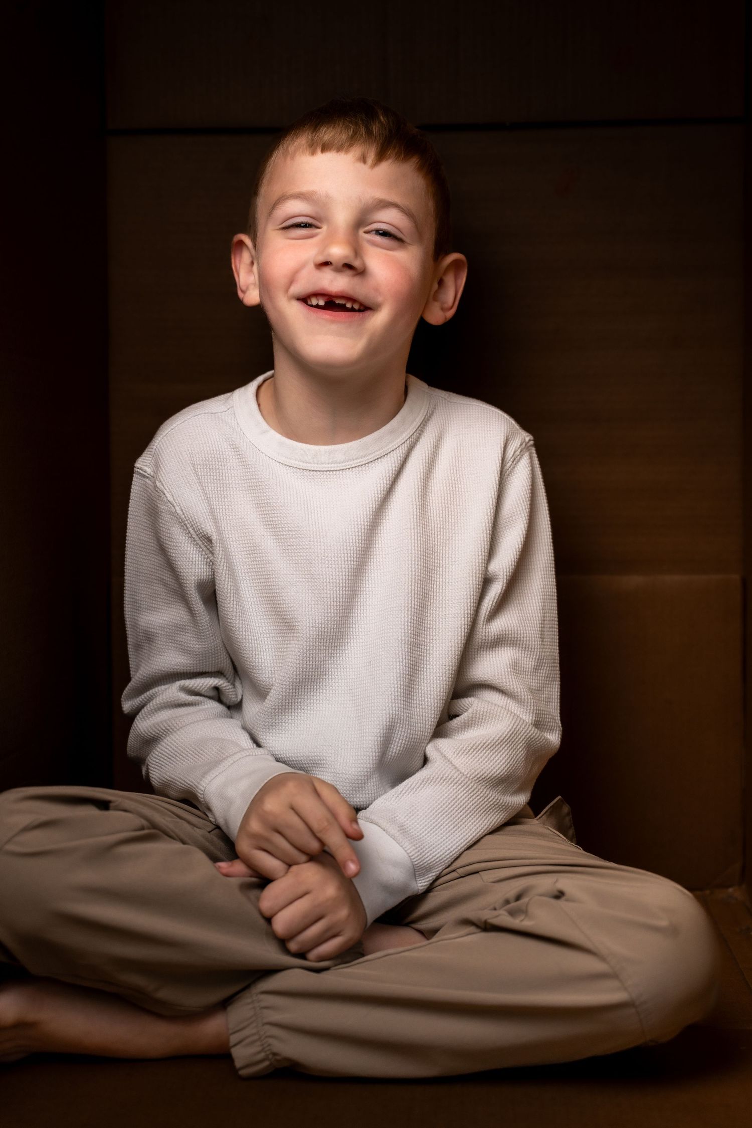 A cheerful young boy with a missing tooth sits cross-legged in a cardboard box, wearing a white sweatshirt and khaki pants.