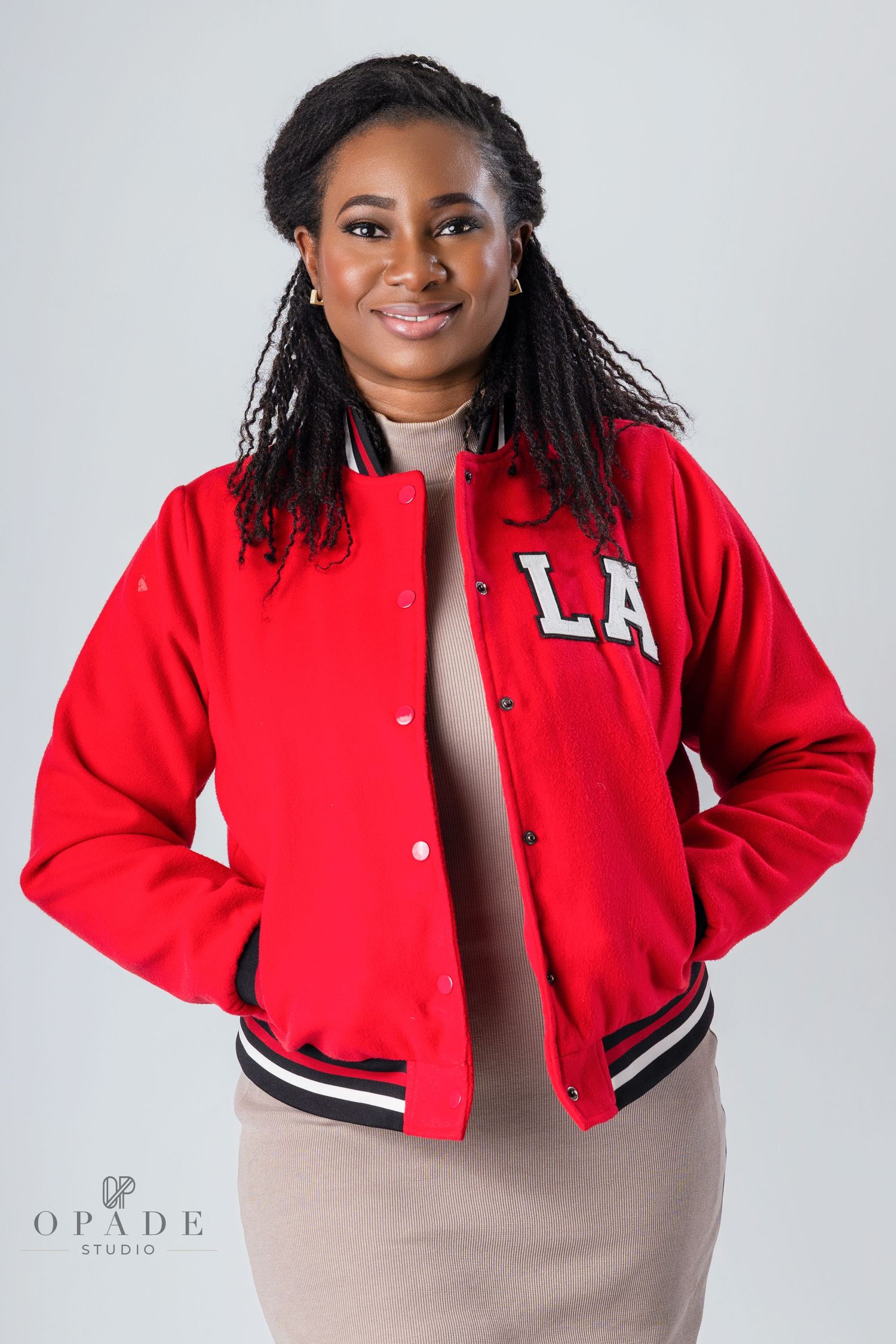 Woman wearing a red varsity jacket standing against a light grey background with hands in pockets.