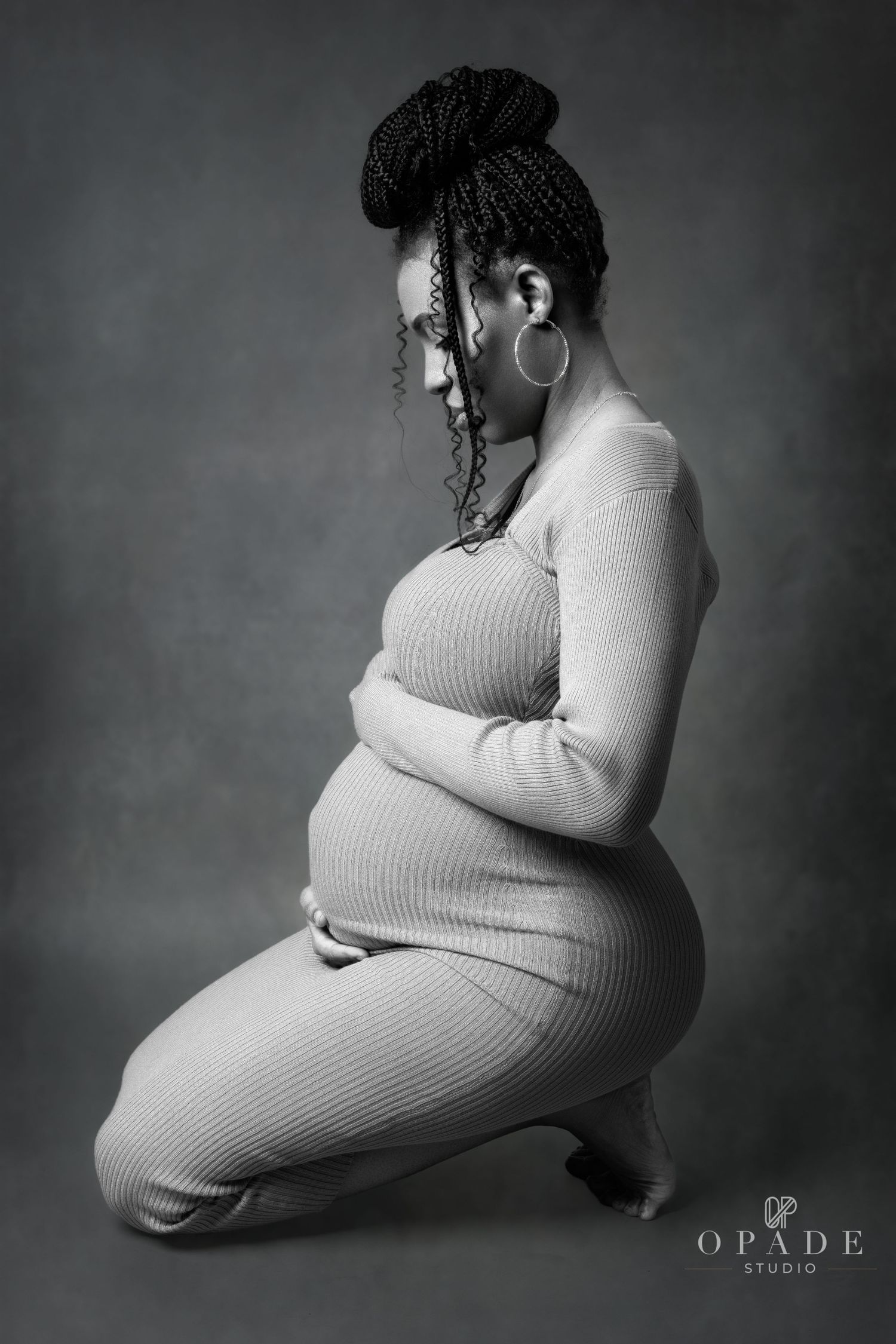 Black-and-white maternity portrait of a pregnant woman kneeling and gently holding her belly in a studio.