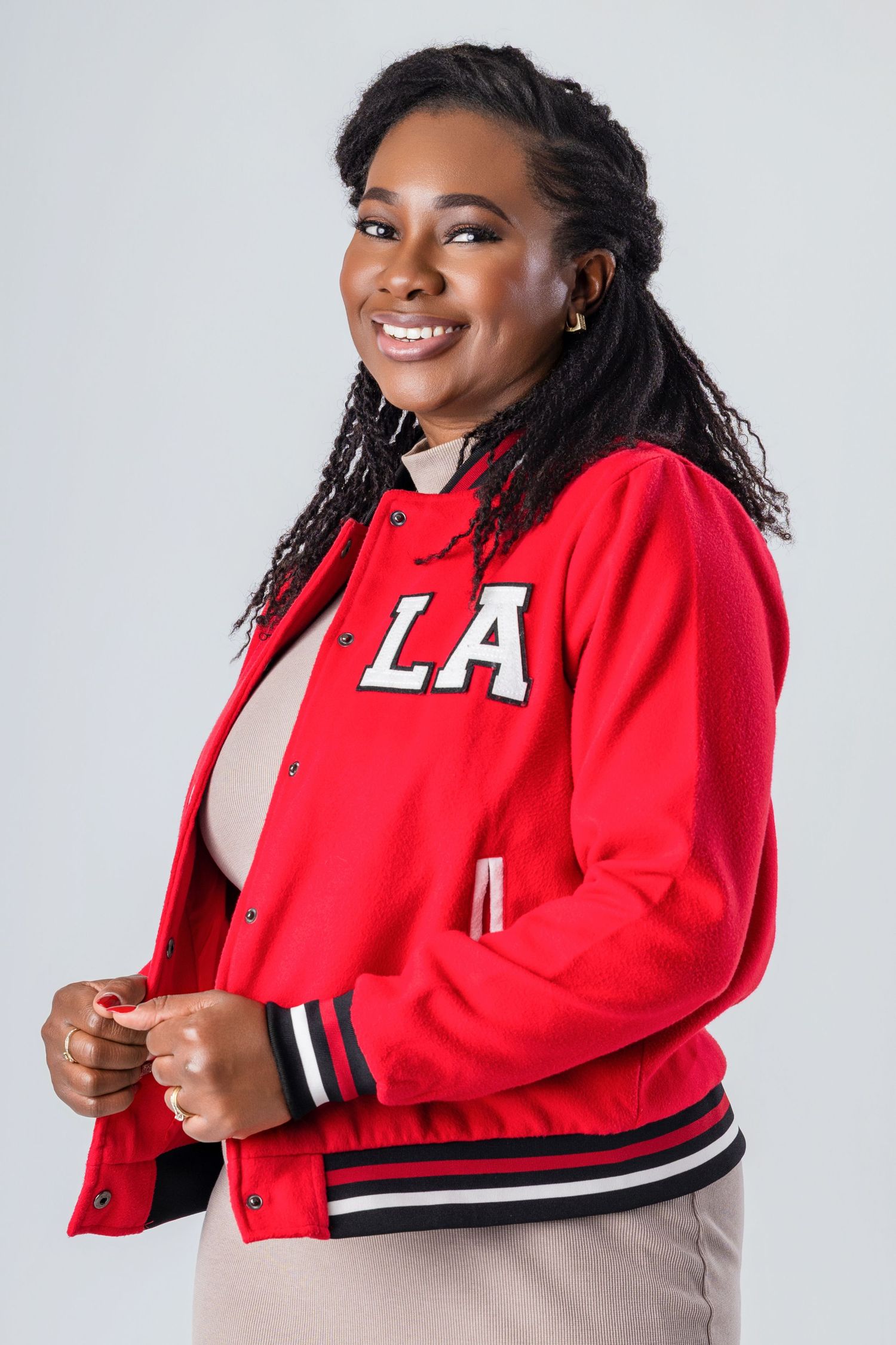 Smiling woman wearing a red varsity jacket with “LA” lettering, posing against a light grey studio background while holding the front of her