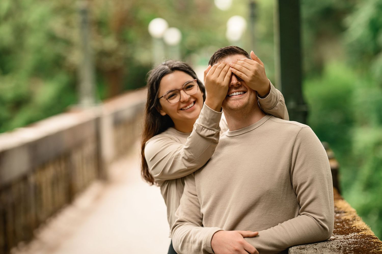 She covers his eyes and they both laugh against the mossy bridge wall, Arboretum proposal photographer, Seattle