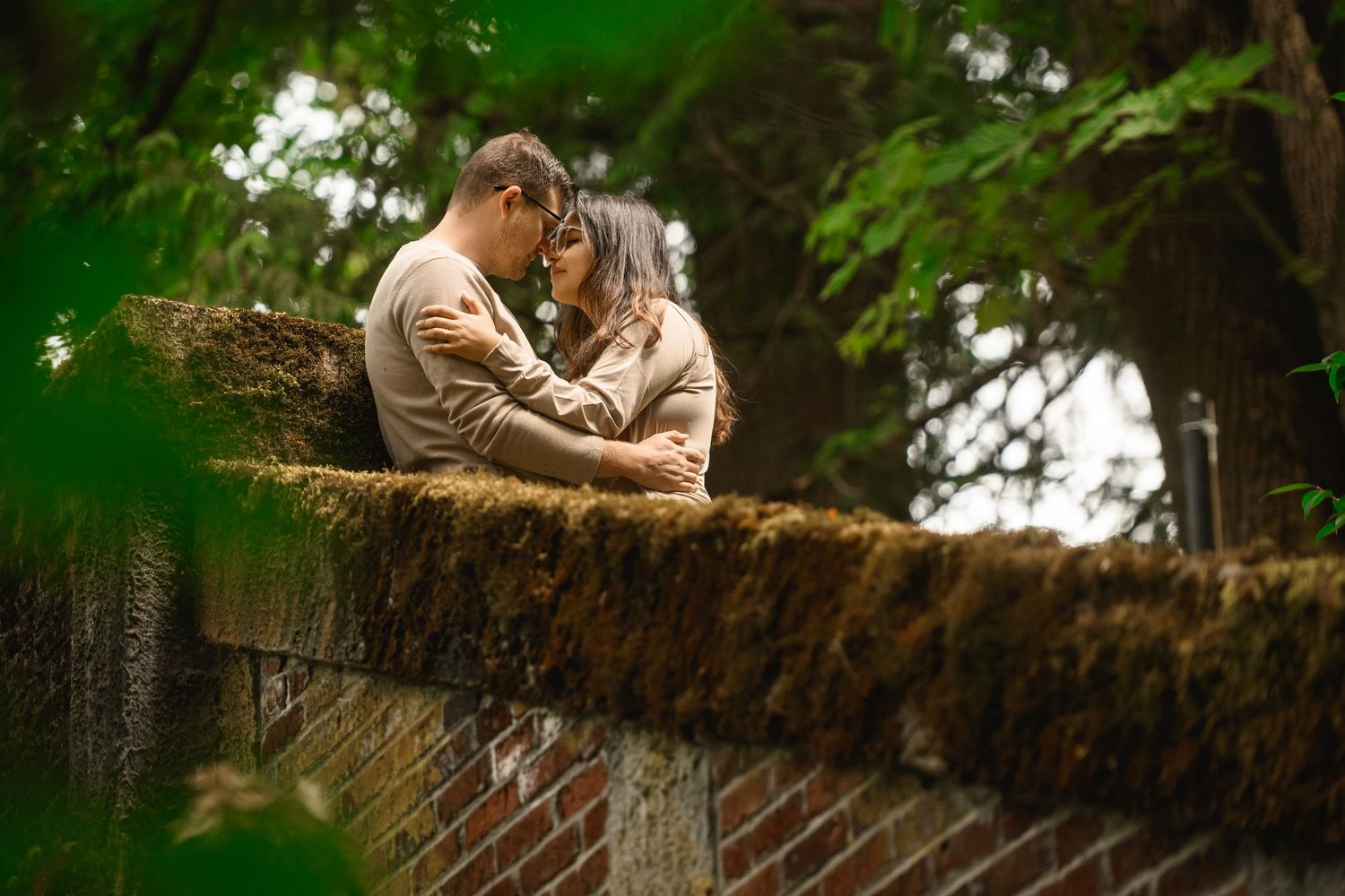Couple close together at a mossy stone wall under green leaves, Arboretum proposal photographer, Seattle