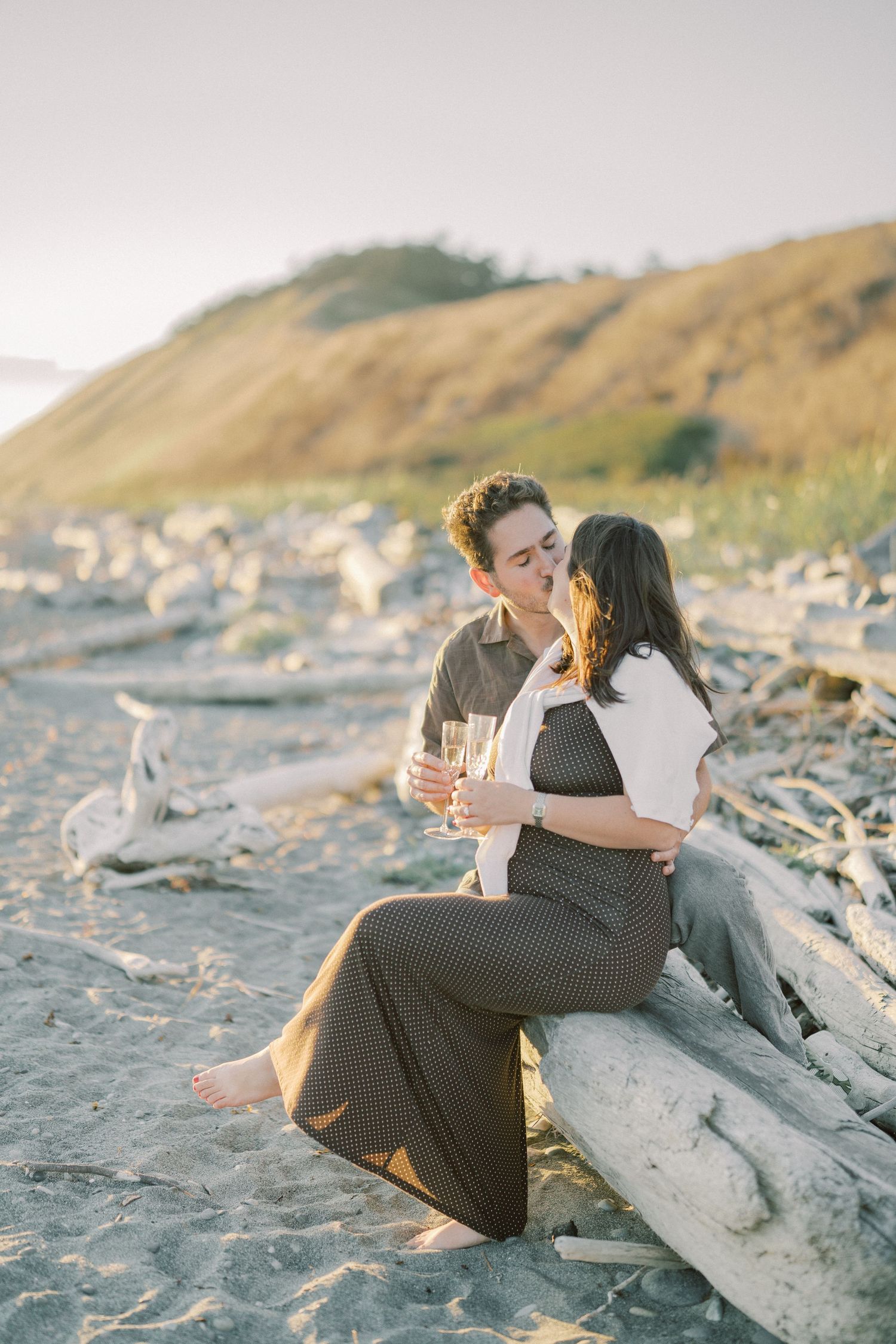Couple sitting on driftwood with champagne below the bluff, Whidbey Island proposal photographer, Whidbey Island