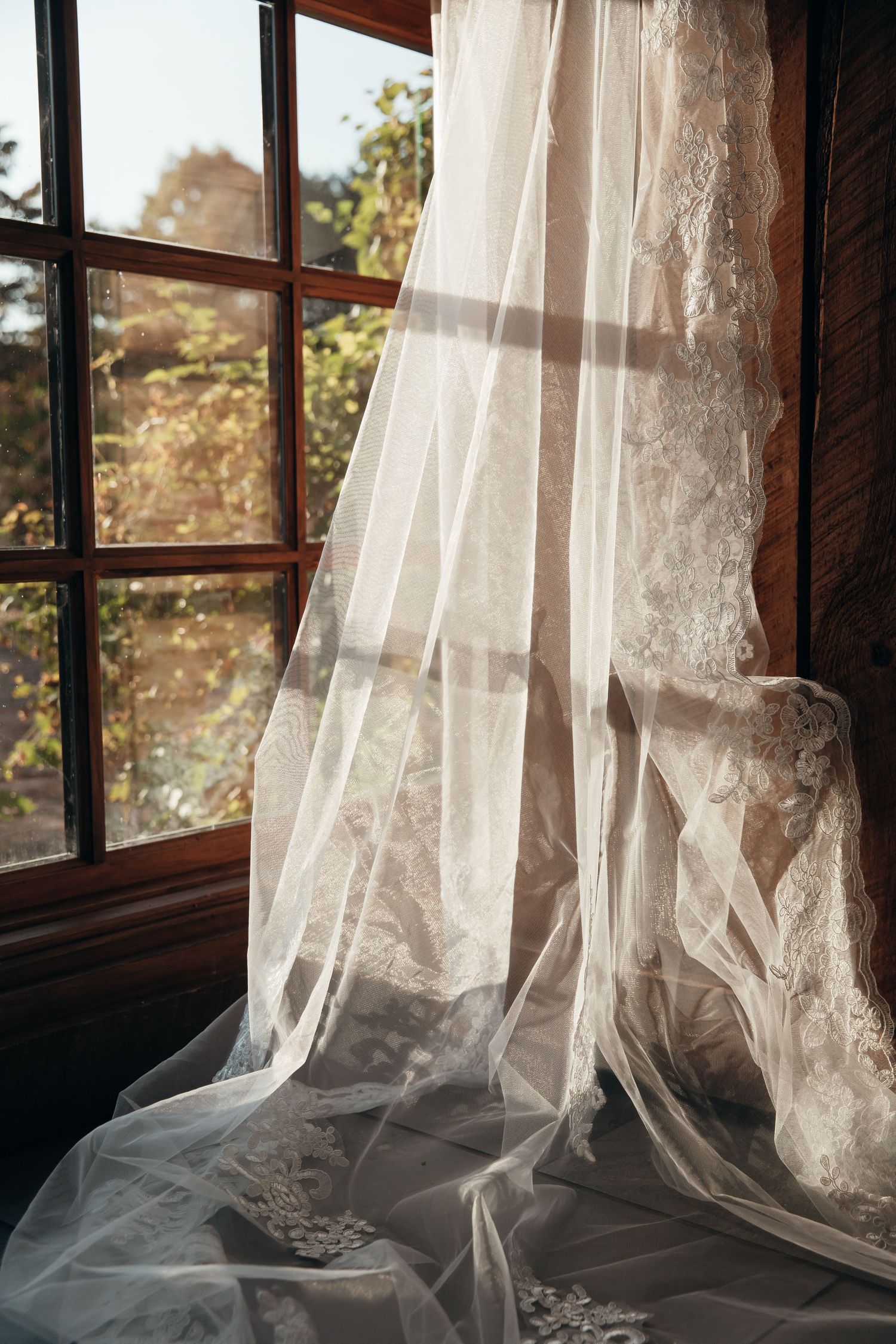 Delicate white lace wedding veil draped by a sunlit window with wooden frames creating a romantic atmosphere.