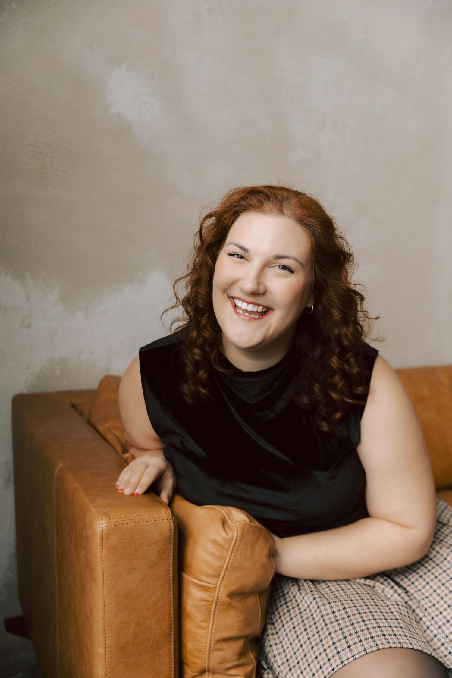 A joyful portrait of someone with curly red hair wearing a black top and sitting beside a tan leather couch.