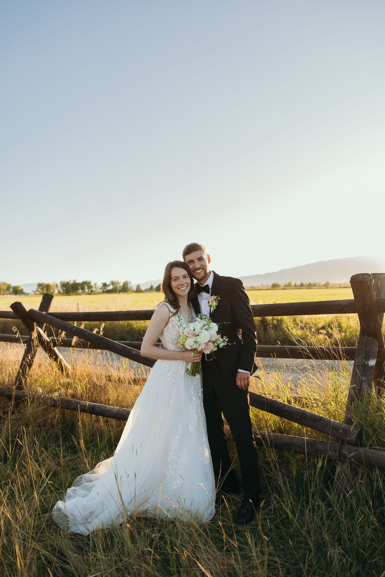 Wedding couple poses near wooden fence in countryside during sunset.