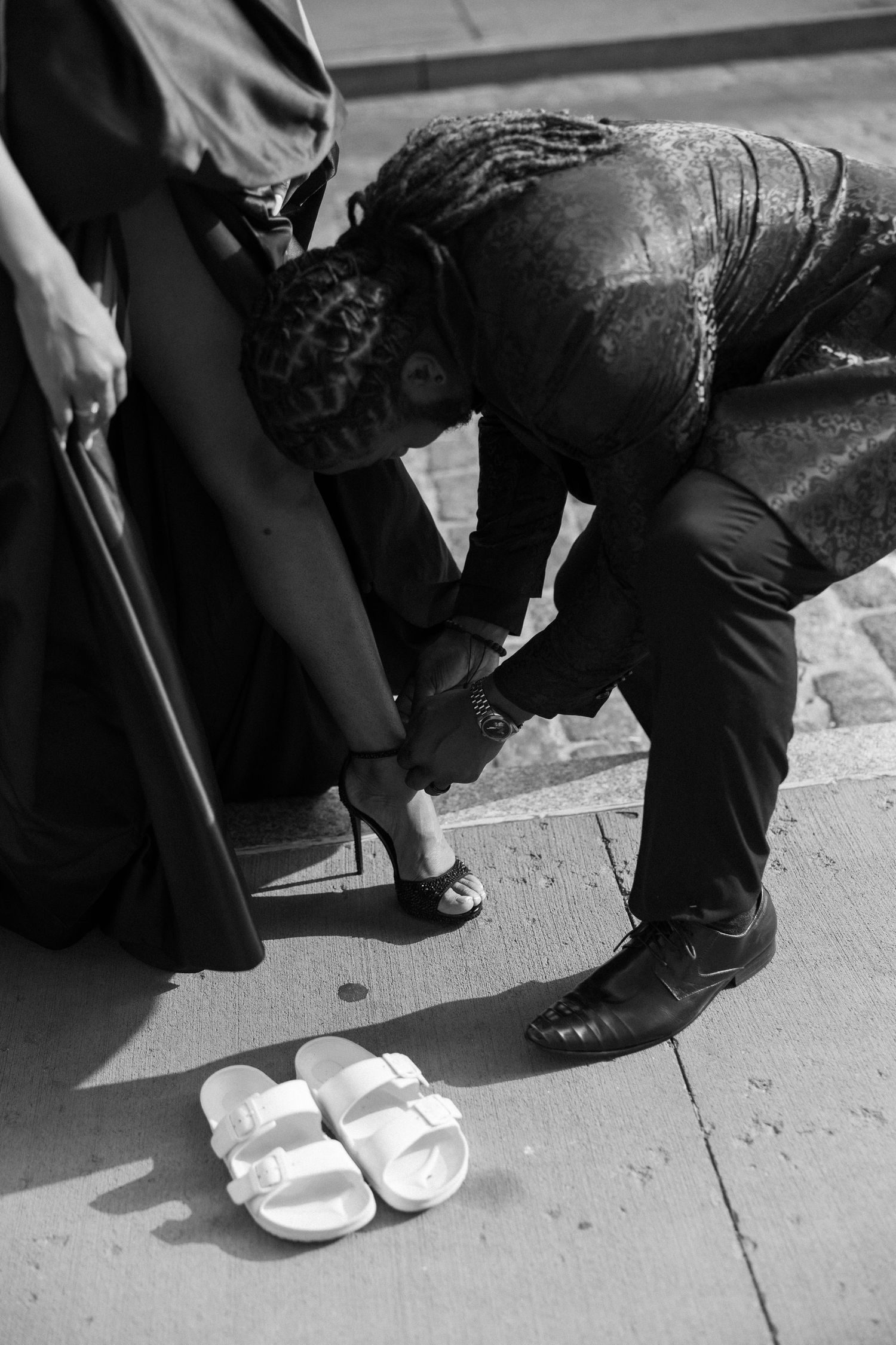 Person in formal wear adjusts their shoes on cobblestone sidewalk.