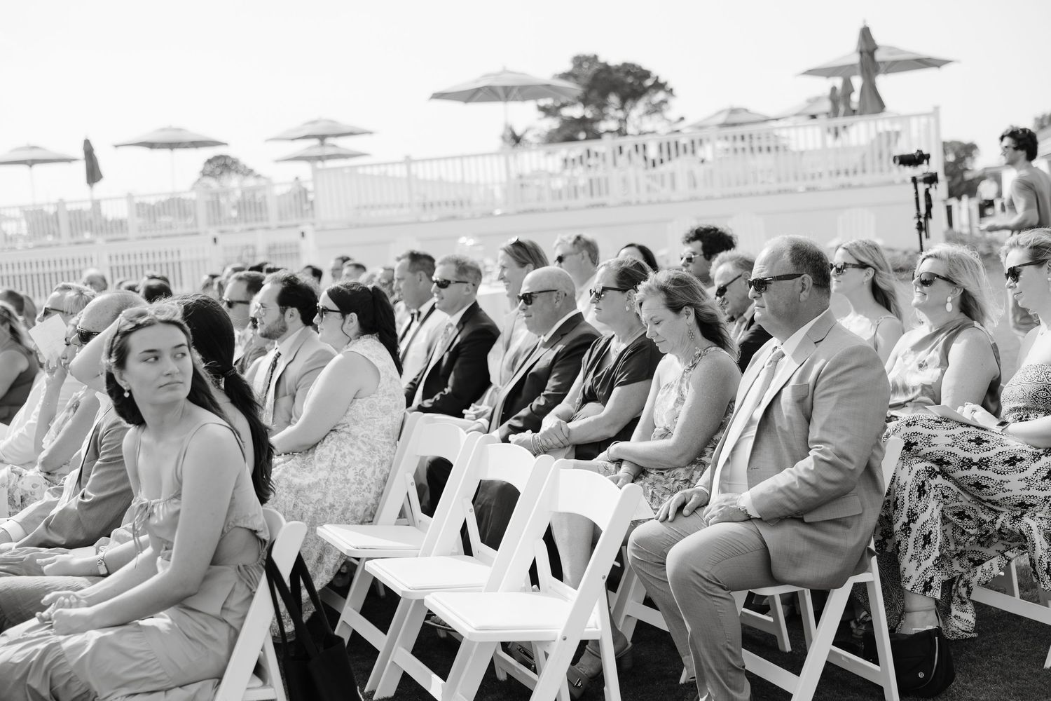 Wedding guests seated in white chairs during an outdoor ceremony.