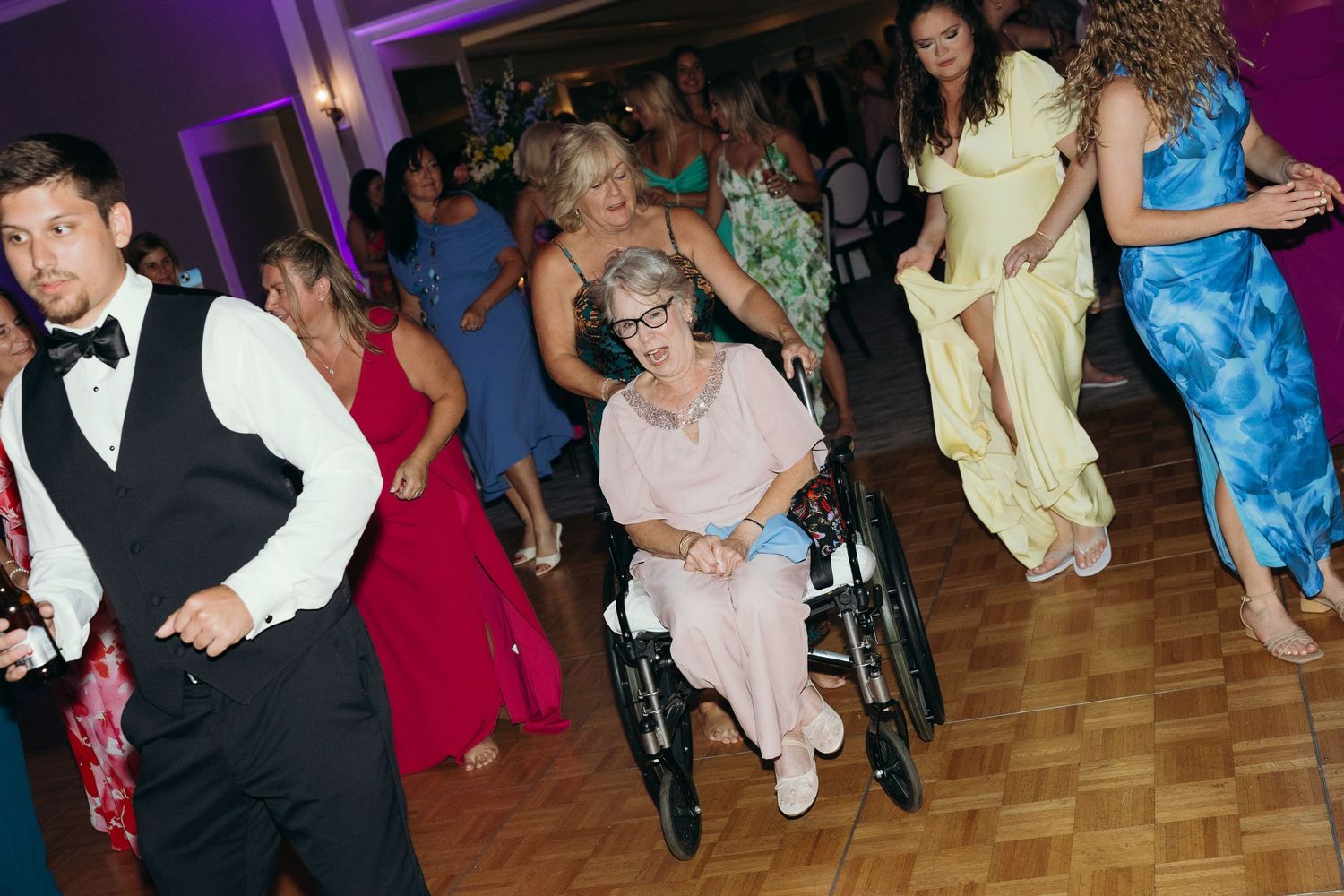 Lively dance floor scene at a wedding reception with colorful dresses and purple ambient lighting.