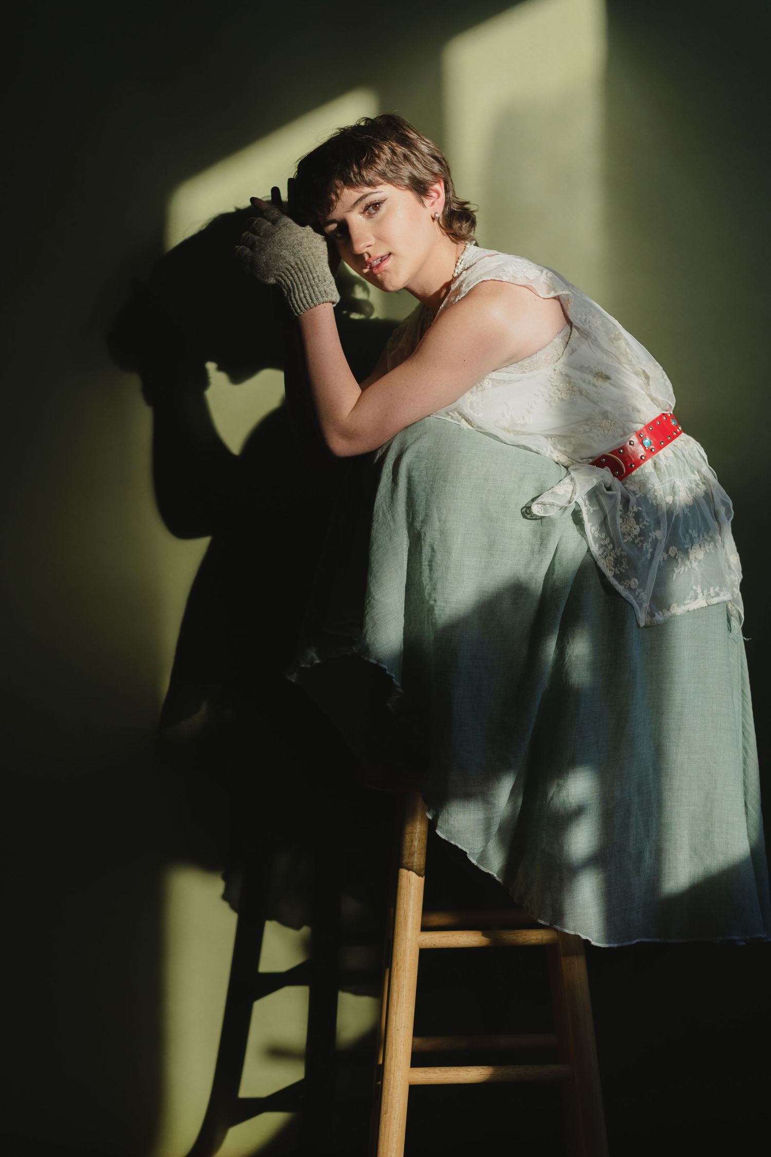 A person in a vintage-style mint green dress and lace gloves poses dramatically against a wall casting shadows.