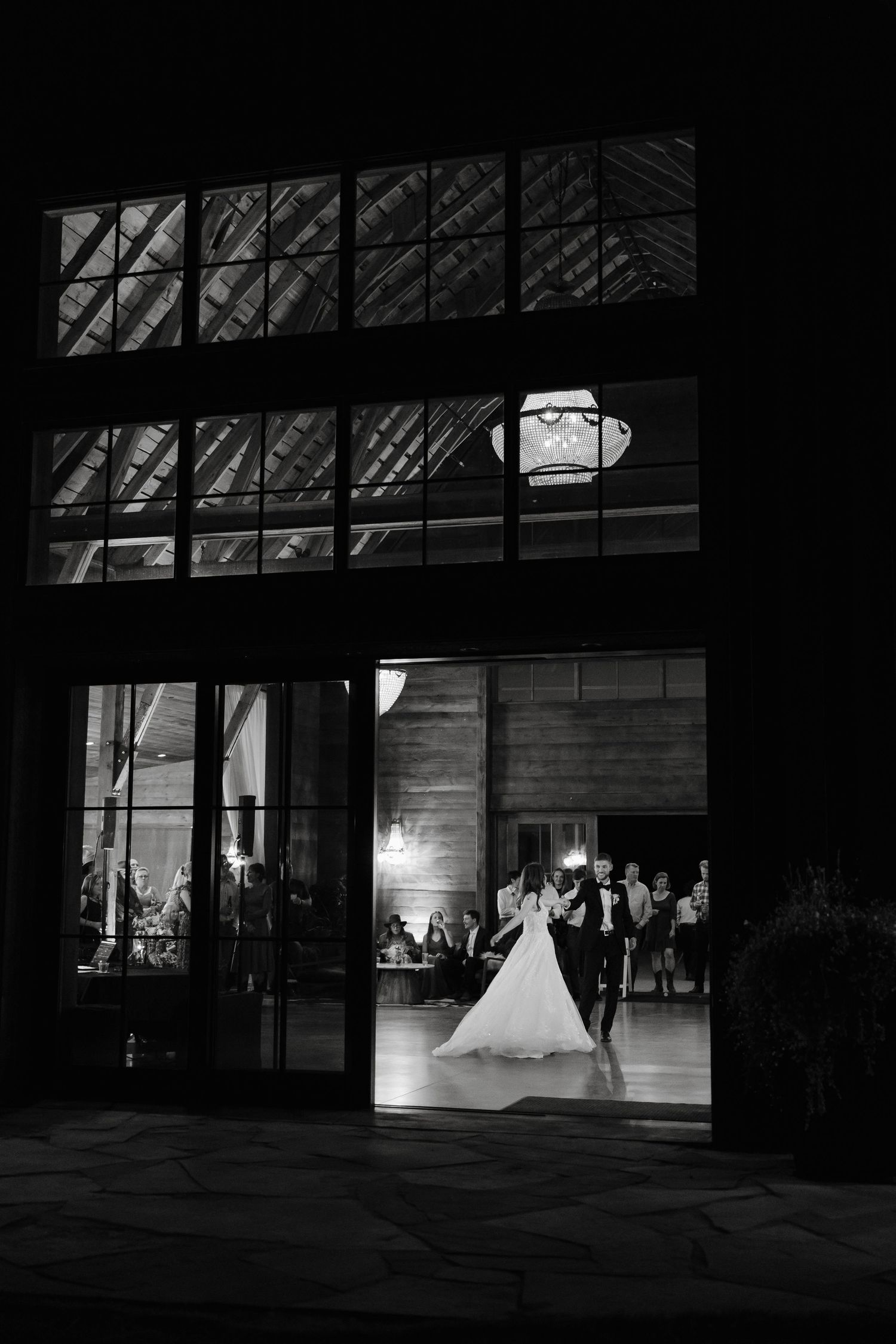 Silhouette of a wedding celebration inside a dark barn venue with dramatic lighting and large windows.
