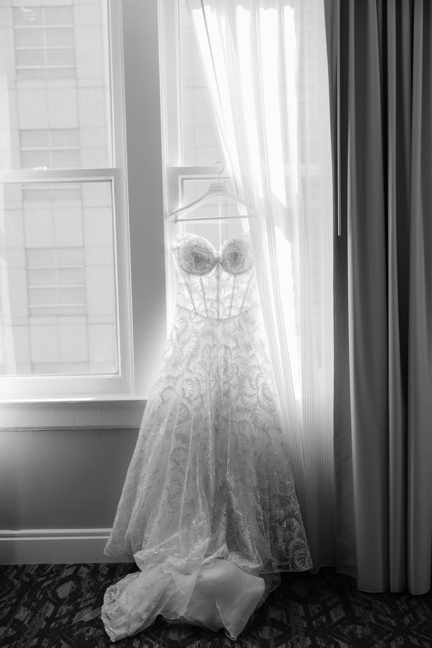 A delicate white lace wedding dress hangs elegantly by a window in black and white photography.