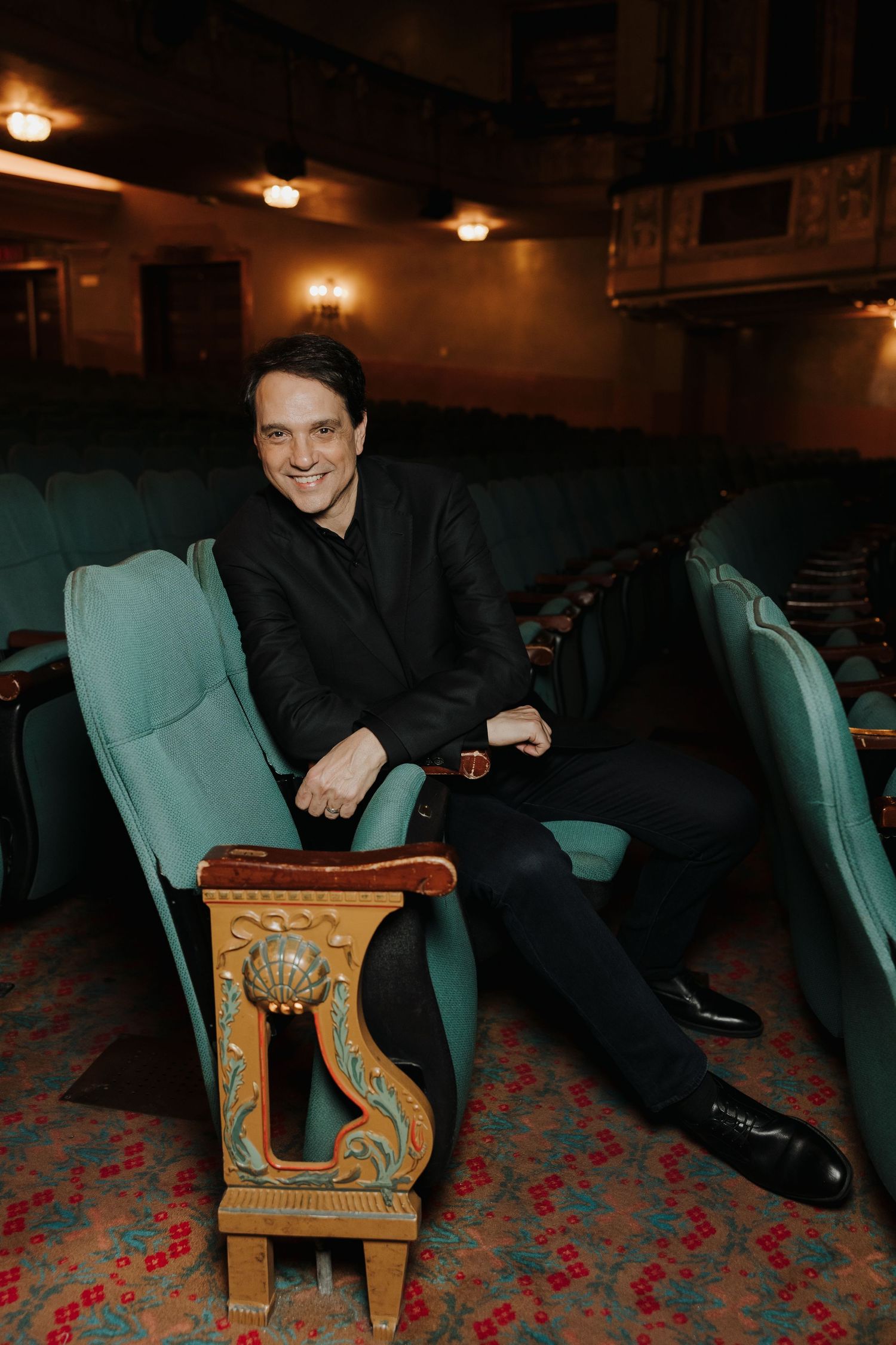 Man in black suit smiling in vintage theater seats with ornate armrests and teal upholstery.