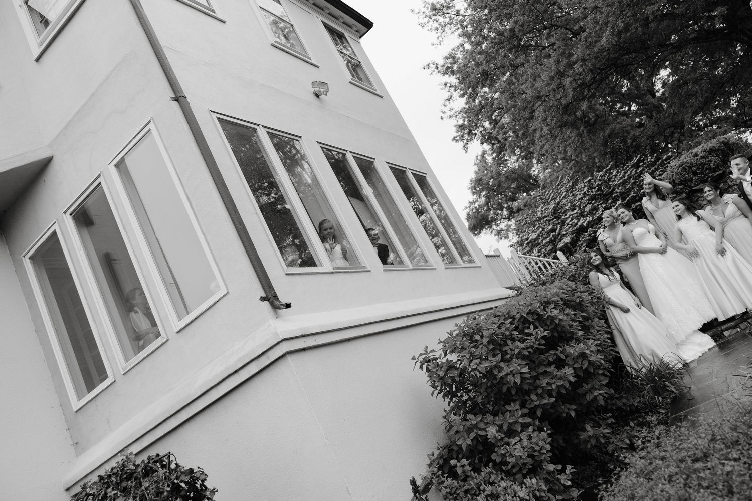 Black and white photo of bridesmaids in white dresses standing outside an elegant building surrounded by lush greenery.