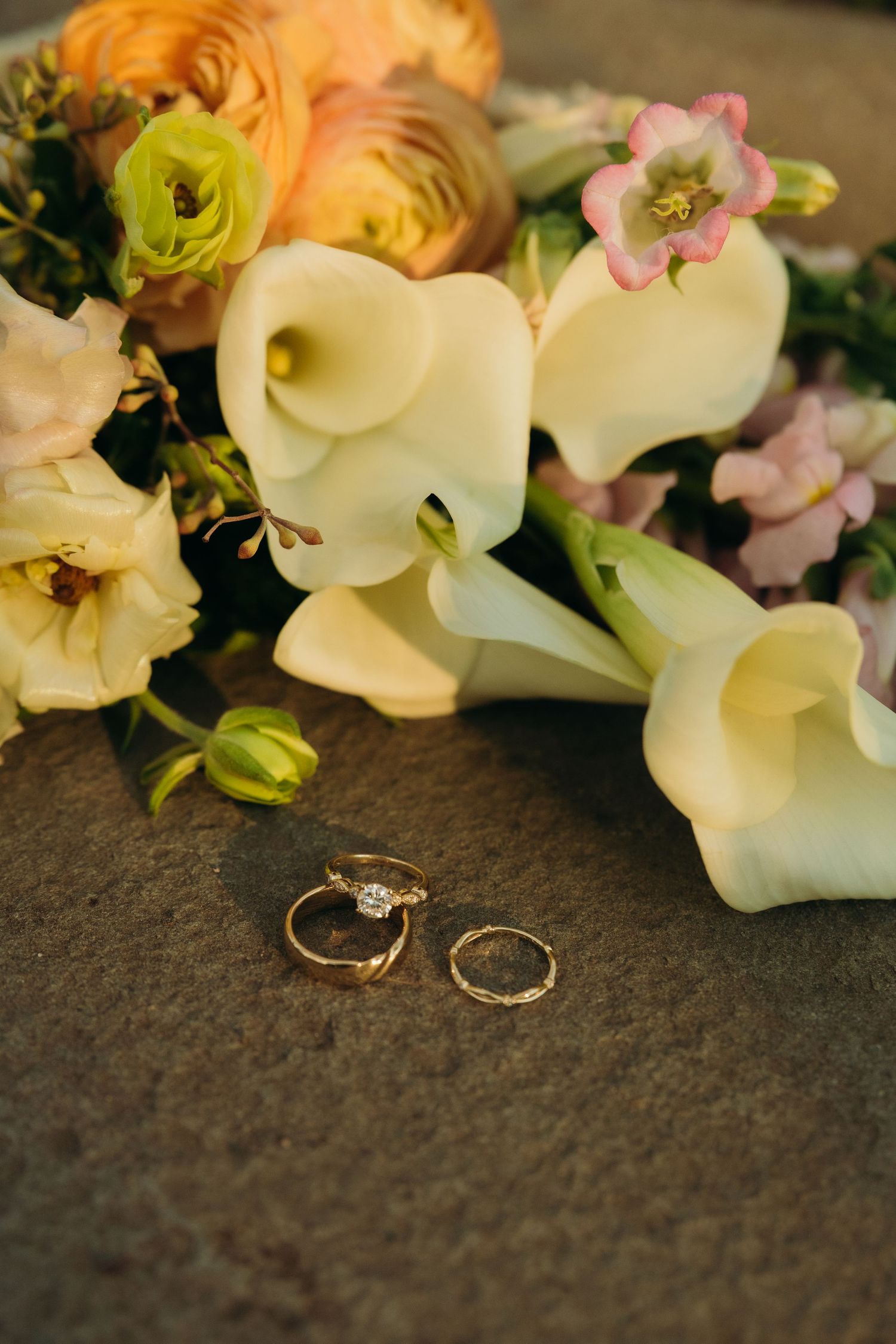 Wedding rings nestled among delicate white calla lilies and pink flowers on dark surface.