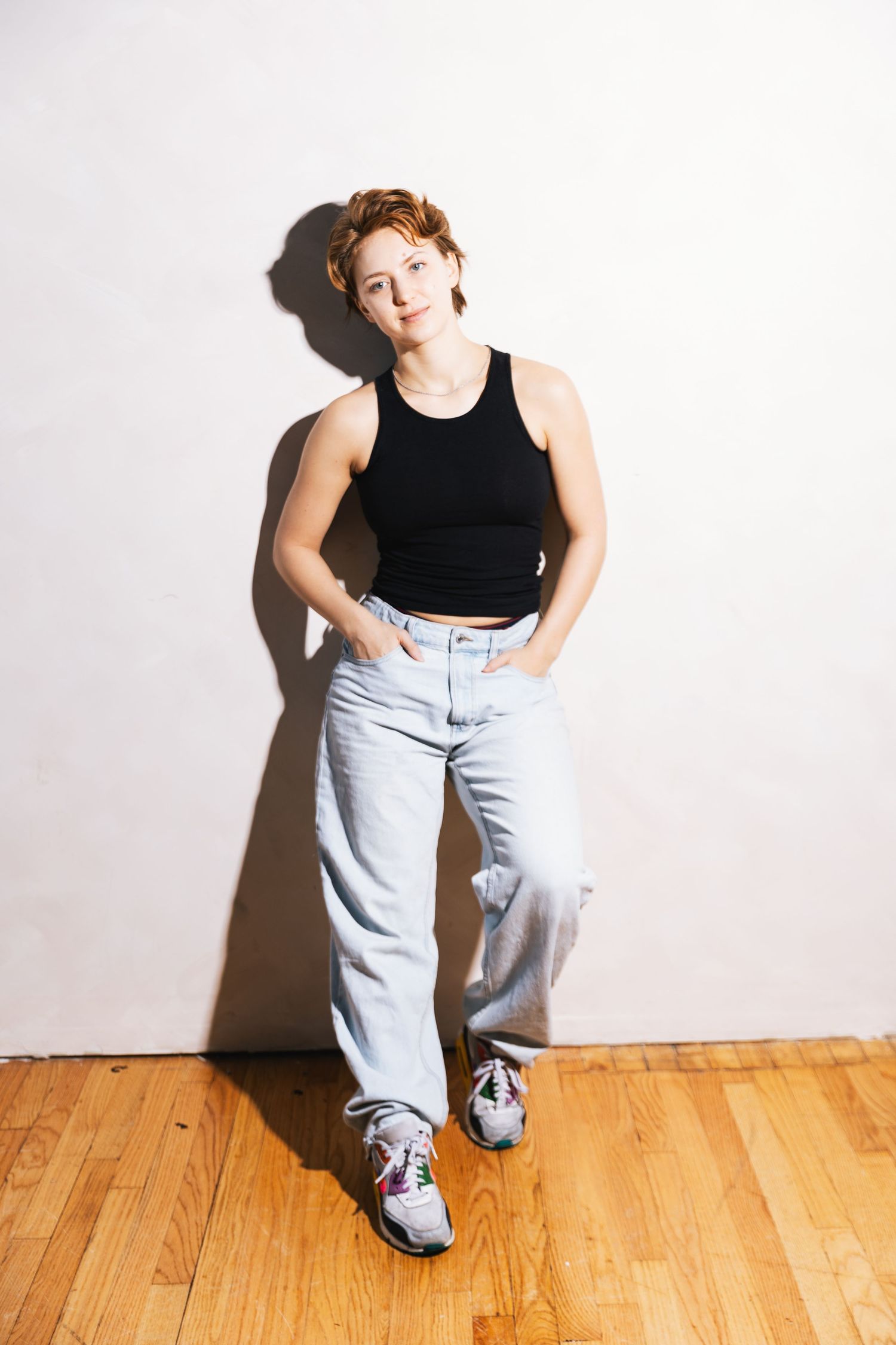 A person in a black tank top and light wash jeans poses casually against a white wall on hardwood floors.