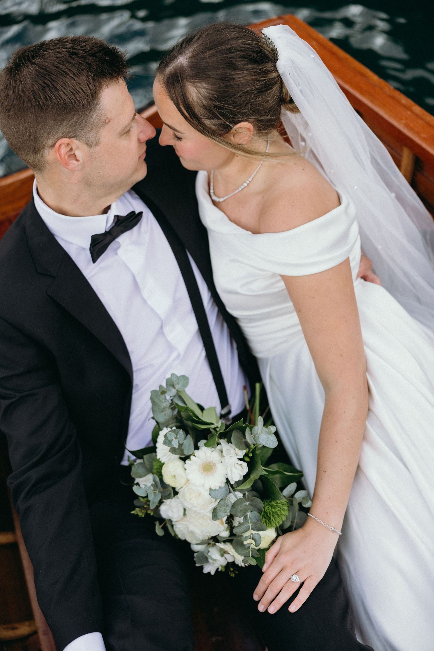 Romantic moment between newlyweds in a wooden boat with white floral bouquet.