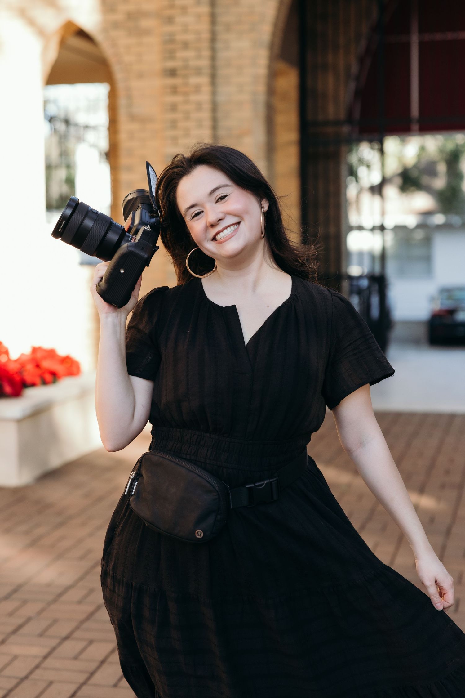 Professional photographer in black dress smiles while holding camera equipment in outdoor brick setting.