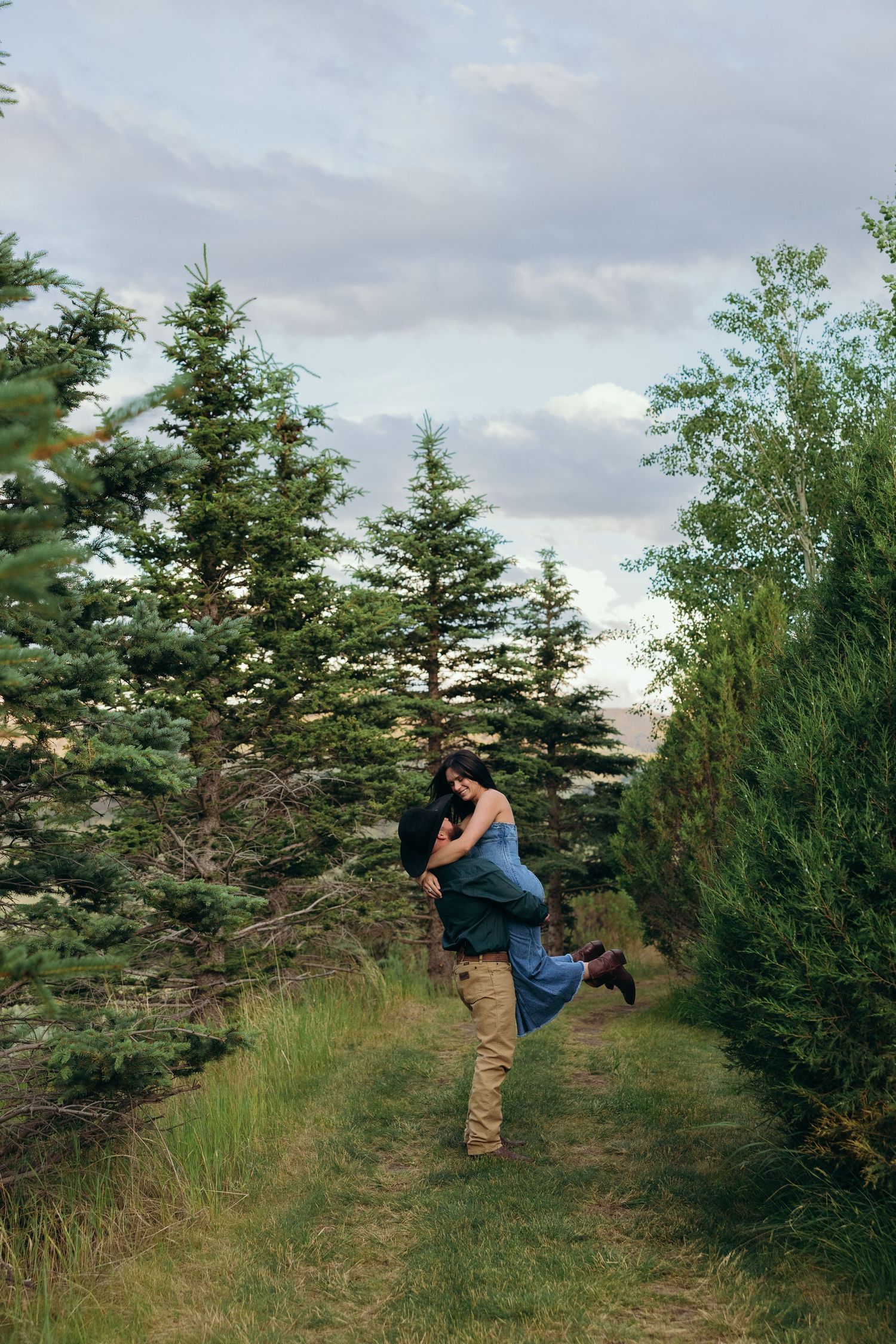 A couple playfully poses between rows of evergreen trees in a rustic outdoor setting.