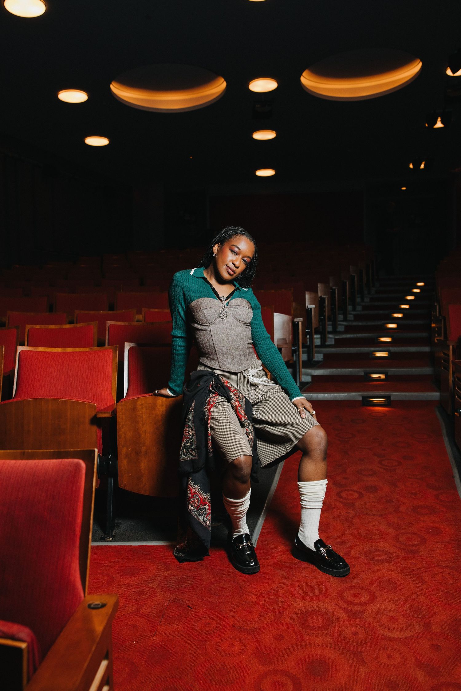 Young woman in teal cardigan and gray corset sits in vintage red theater seats on patterned carpet.