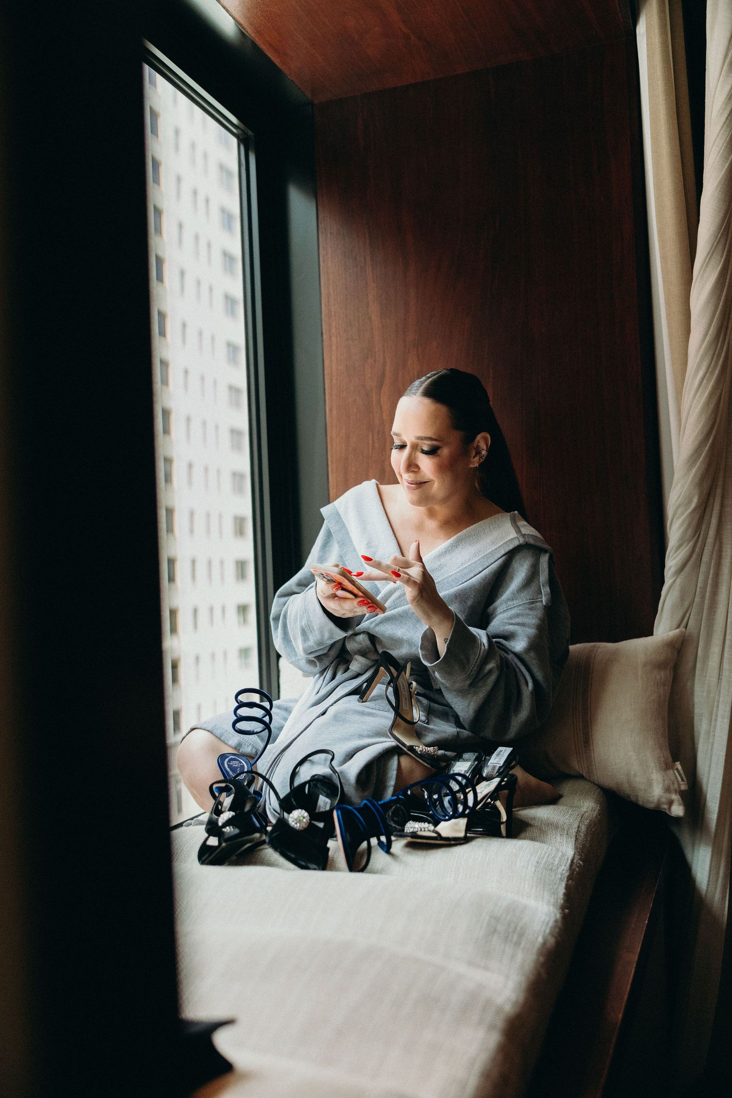 Person in a cozy bathrobe sits by window looking at phone while relaxing on cushioned windowsill.
