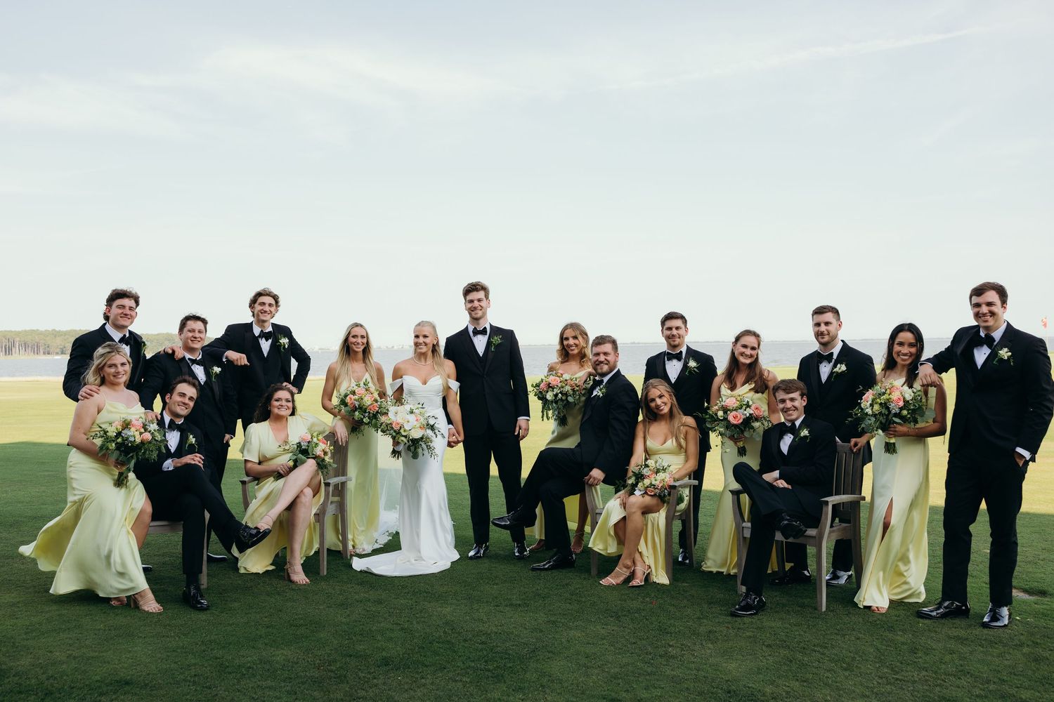 Wedding party group photo with bridesmaids in light dresses and groomsmen in dark suits on a grassy hill.