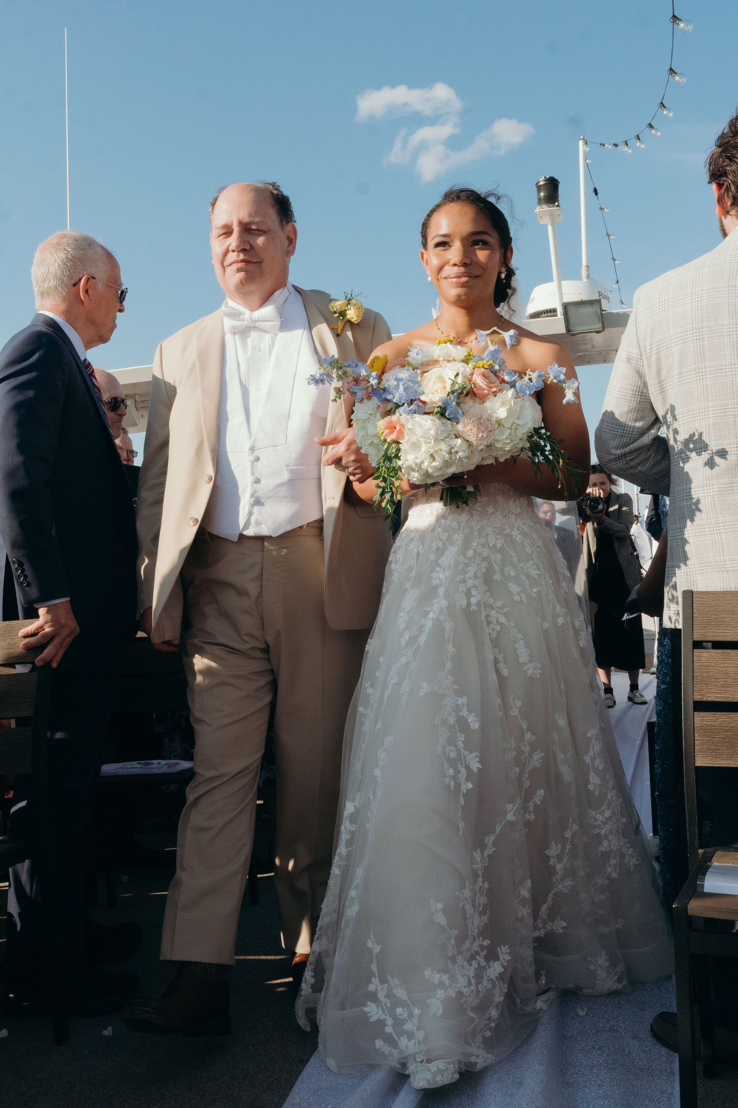 Wedding guests and bridal party gather on a sunny outdoor deck with blue skies.