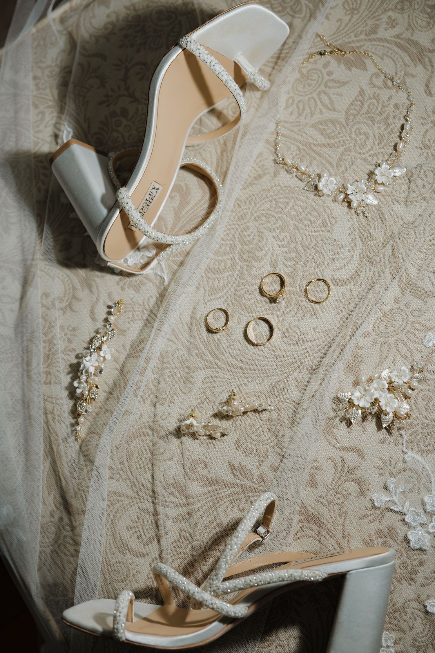 Elegant bridal accessories flat lay featuring pearl heels, gold rings, floral earrings, and a delicate necklace on lace fabric.