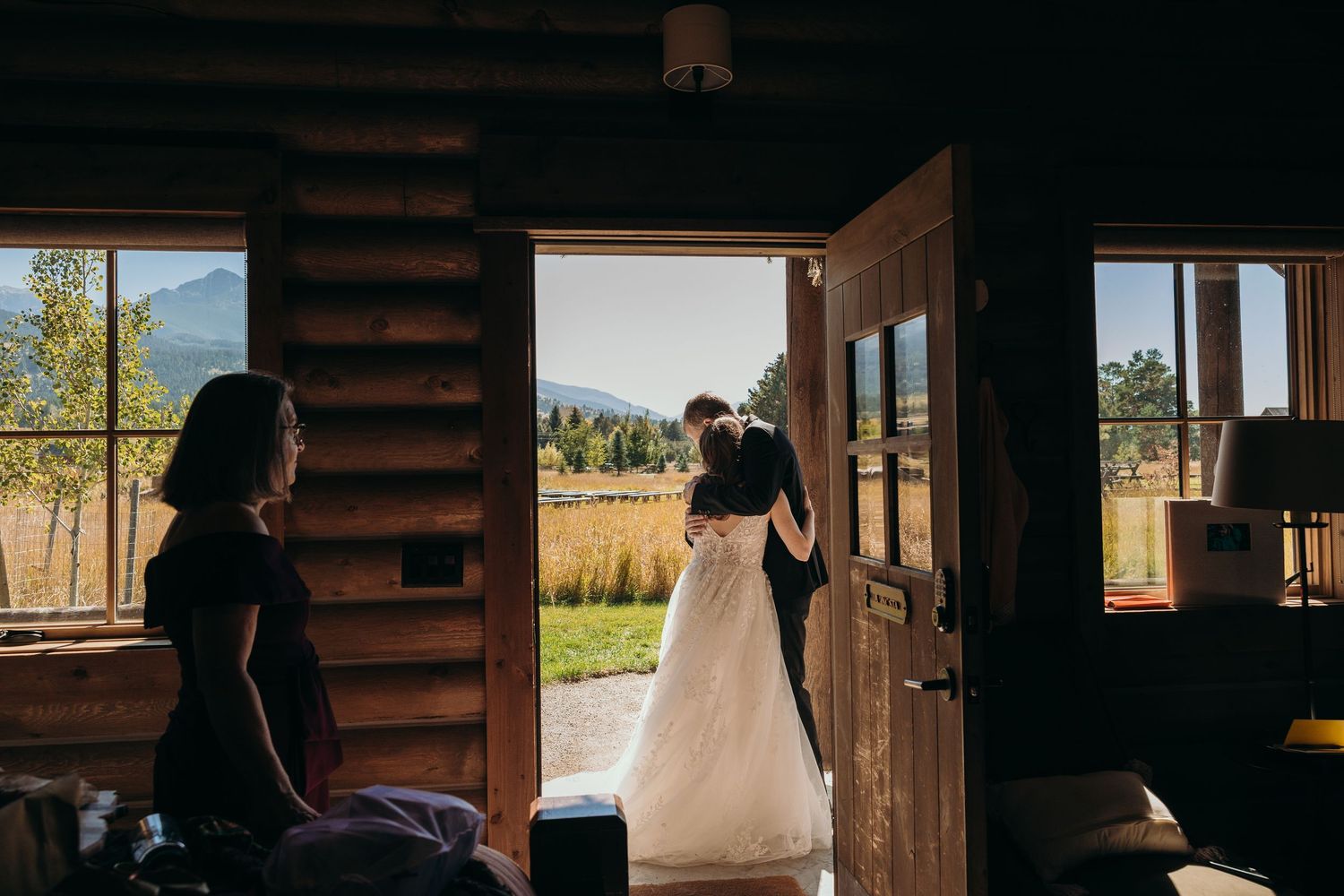 Newlyweds stand in silhouette inside a rustic wooden cabin with sunlight streaming through.
