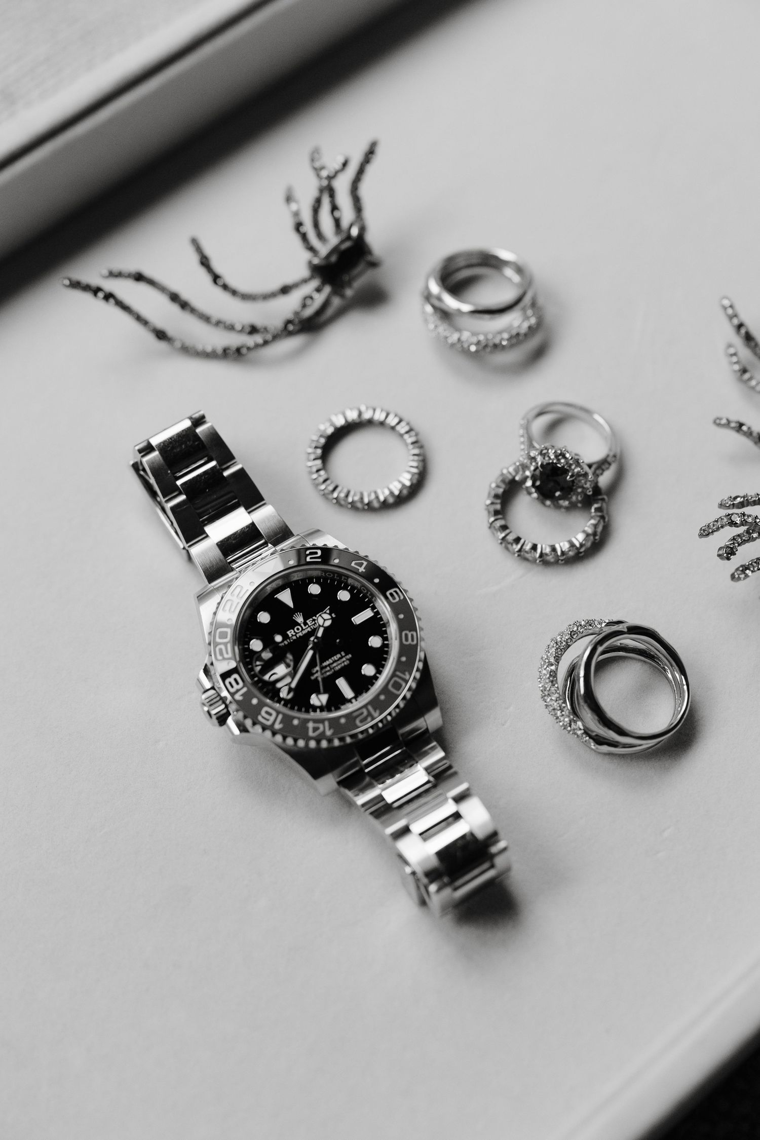 Silver watch and jewelry pieces arranged on a light gray surface in black and white photography.