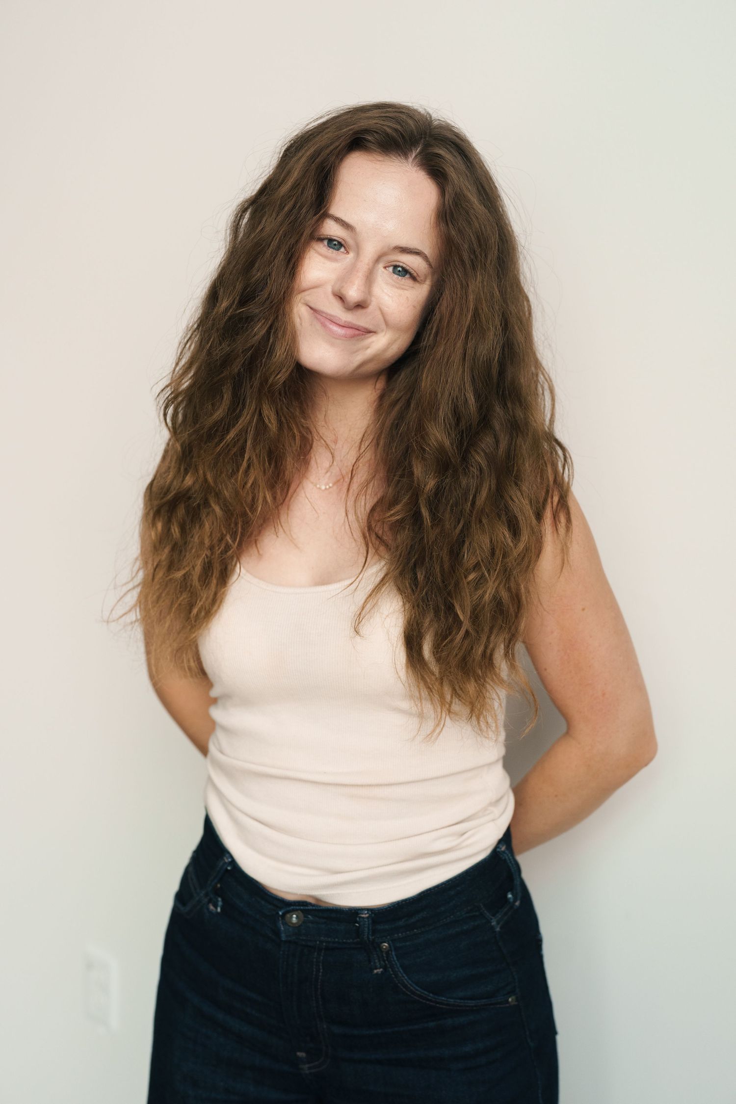 Young woman with long wavy brown hair smiling, wearing a cream tank top and dark jeans against a white wall.